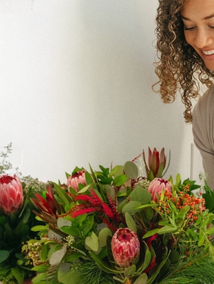 A woman smiles while arranging a vibrant bouquet with striking protea flowers and greenery.