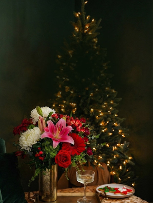 Elegant holiday floral arrangement with red roses, lilies, and festive greenery beside a decorated tree.