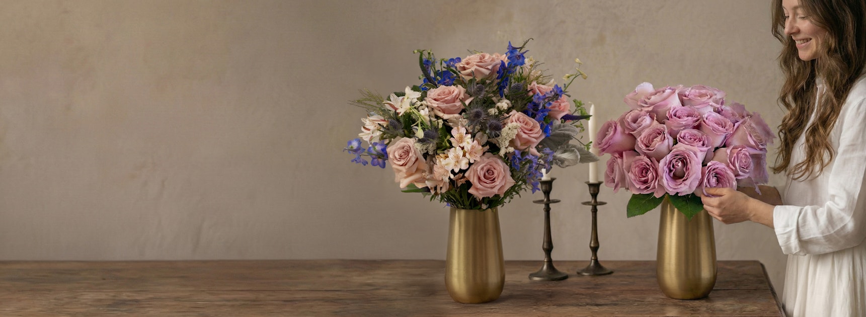 A woman arranges pink and lavender roses in elegant golden vases on a wooden table.