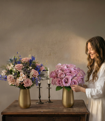 A cheerful woman arranging elegant pastel roses in stylish gold vases on a wooden table.