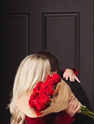 A couple shares a tender kiss while holding a stunning bouquet of red roses.