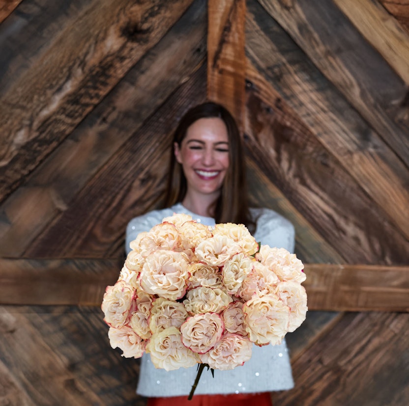 A joyful woman presenting a lush bouquet of soft peach roses against a rustic backdrop.