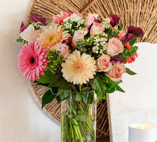 A vibrant floral arrangement featuring pink gerbera daisies, roses, and lush greenery in a glass vase.