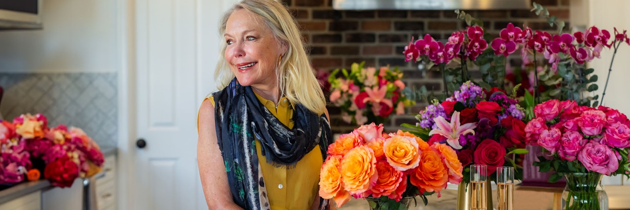 A cheerful woman holds a vibrant bouquet of roses in a beautifully arranged kitchen filled with flowers.