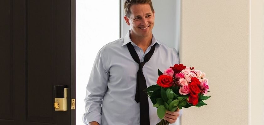 A smiling man in a tie holding a vibrant bouquet of red and pink roses at the door.