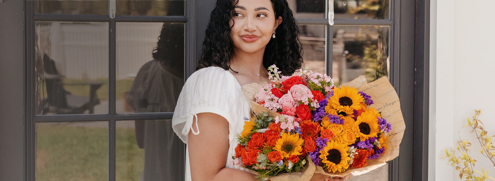 A woman holds a vibrant bouquet of sunflowers and mixed flowers outdoors, smiling cheerfully.