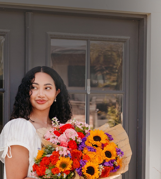 Smiling woman holding a vibrant flower bouquet with sunflowers and roses, standing by a window.