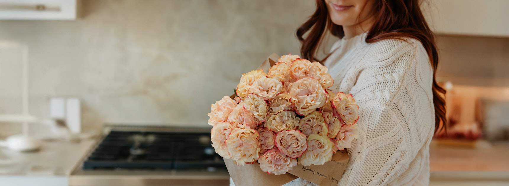 A woman holding a beautiful bouquet of soft peach roses in a cozy kitchen setting.