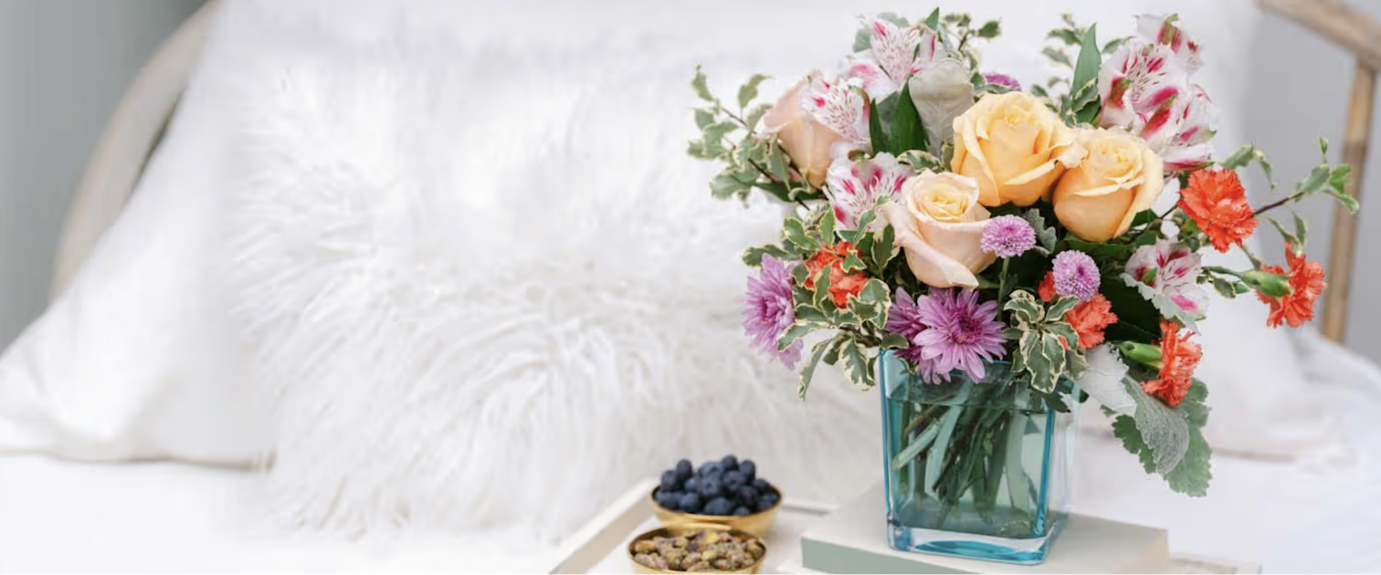 Cheerful floral arrangement of peach roses, bright blooms, and greenery on a cozy bed.