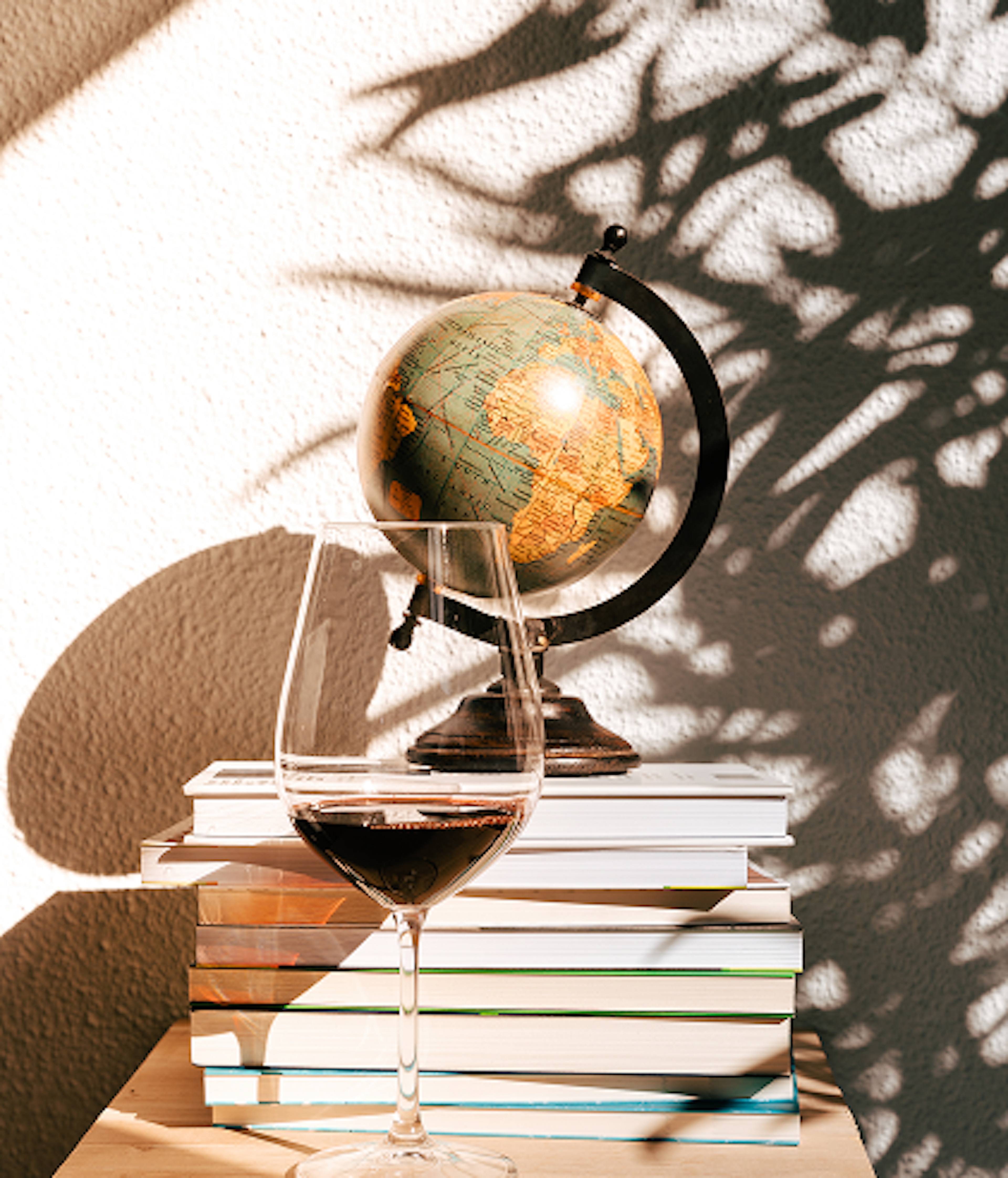 Um copo de vinho tinto está em primeiro plano, ladeado por uma pilha de livros e um globo terrestre, com sombras projetadas de folhagem na parede de fundo
