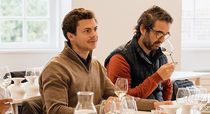 Dois homens estão sentados a uma mesa a participar numa prova de vinhos, com um deles a cheirar o vinho e o outro a observar