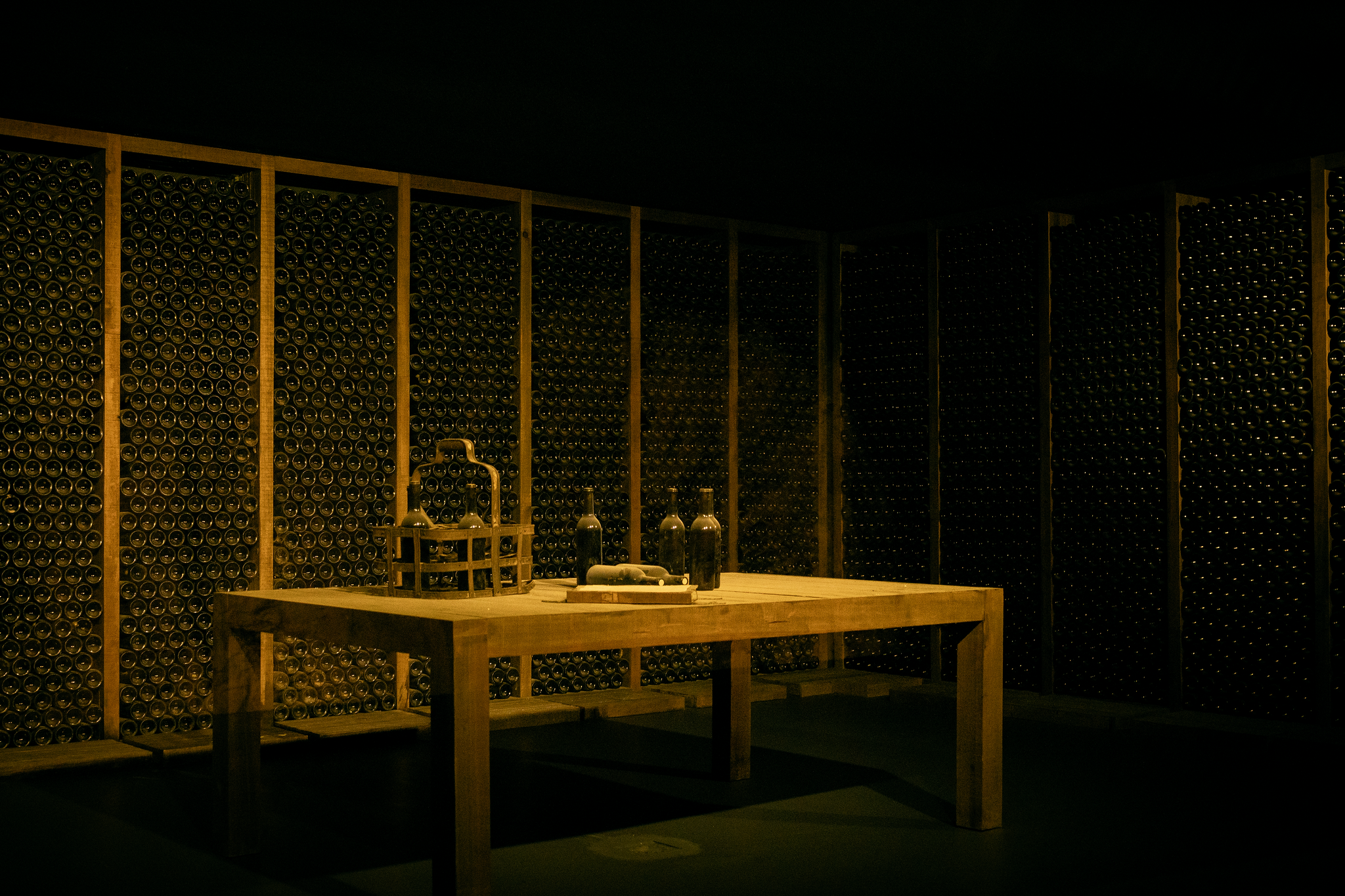 Uma adega escura apresenta paredes repletas de garrafas de vinho e uma mesa de madeira ao centro com mais garrafas e um cesto de transporte