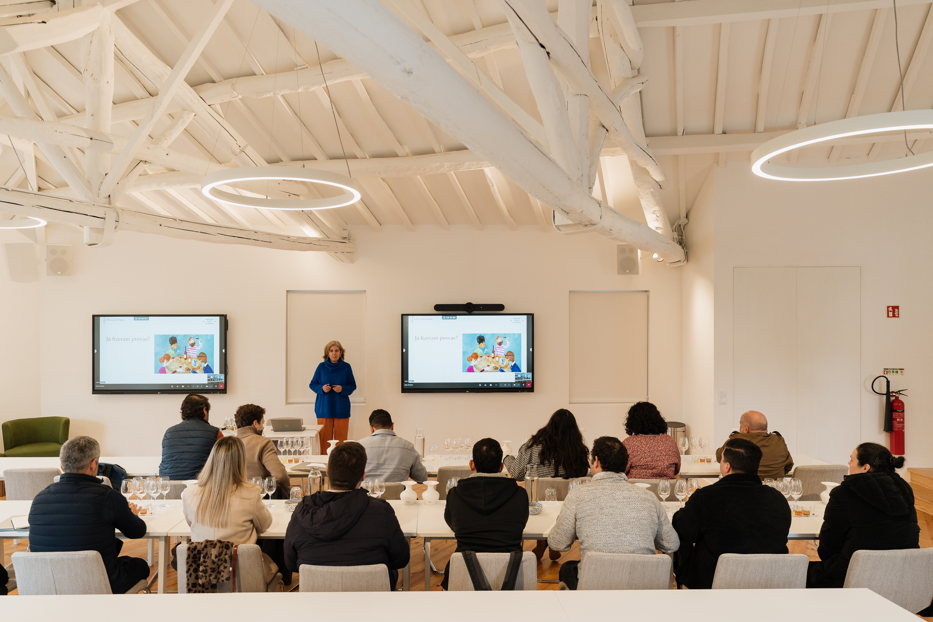 Uma oradora faz uma apresentação em frente a dois ecrãs a um grupo de pessoas sentadas em longas mesas com copos para degustação, numa sala de formação iluminada