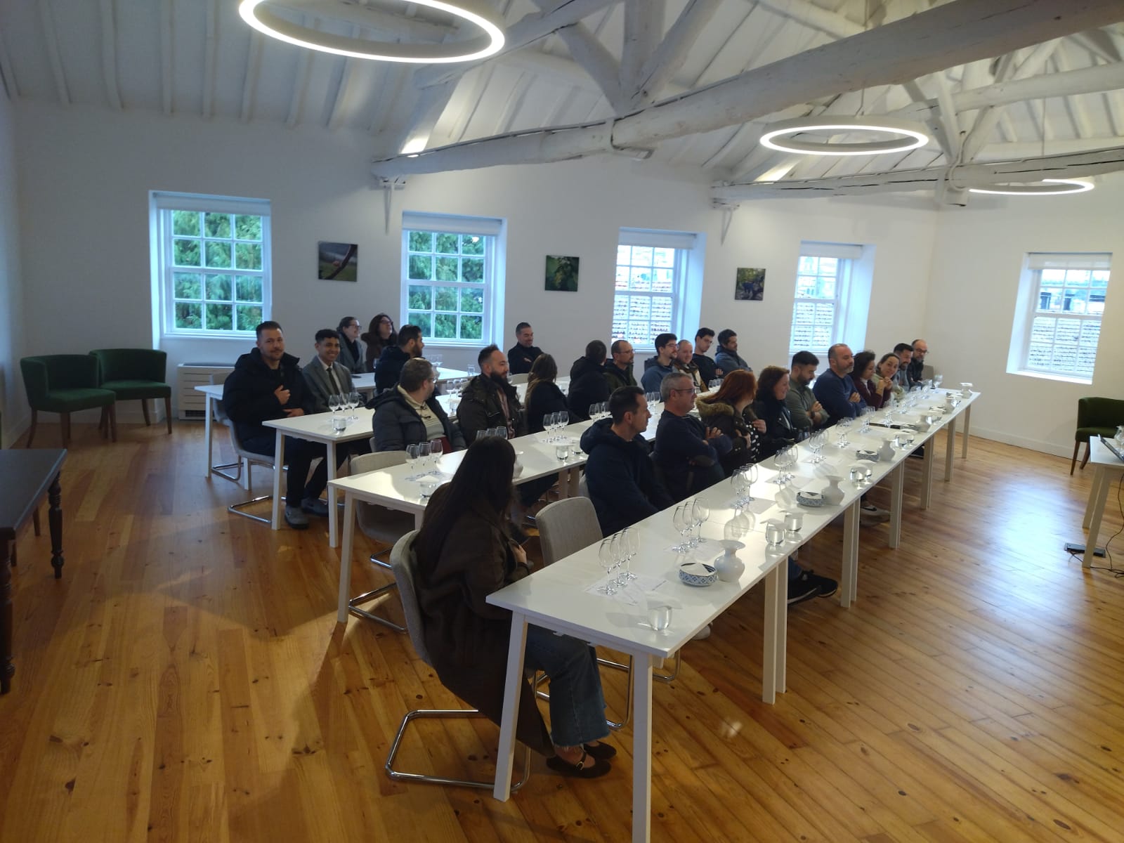 Um grupo de pessoas está sentado em mesas longas com copos de degustação, participando numa aula ou prova numa sala iluminada com teto de madeira