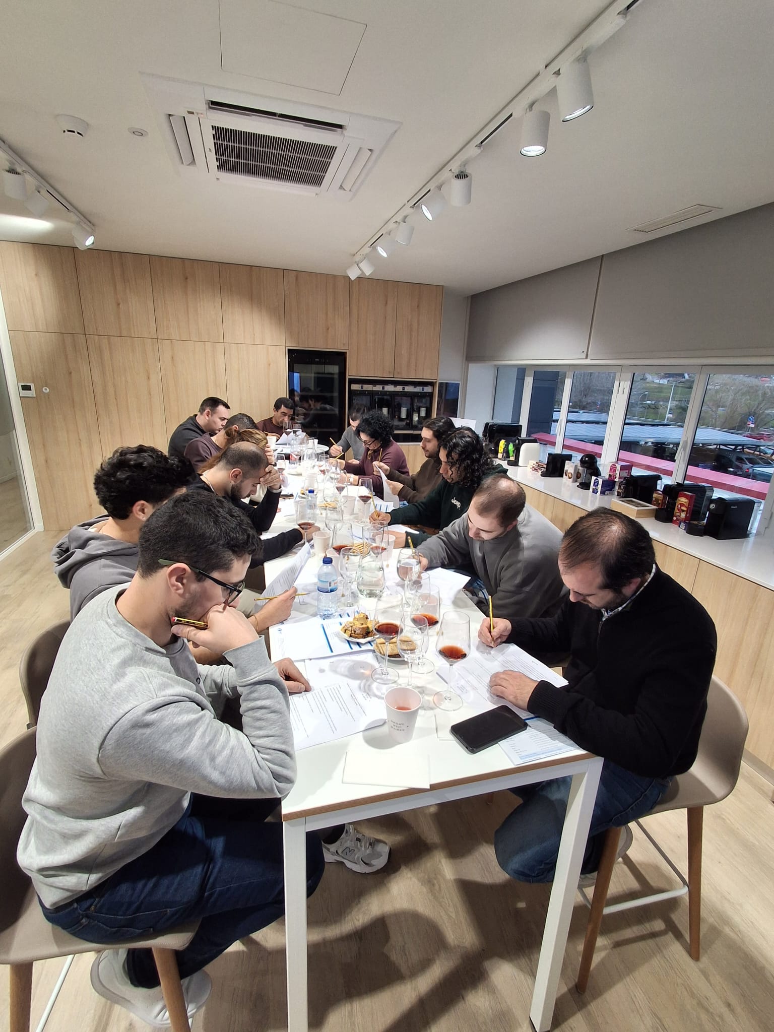 Um grupo de pessoas está sentado à volta de mesas a fazer anotações, com copos de degustação e alguns petiscos, num ambiente moderno