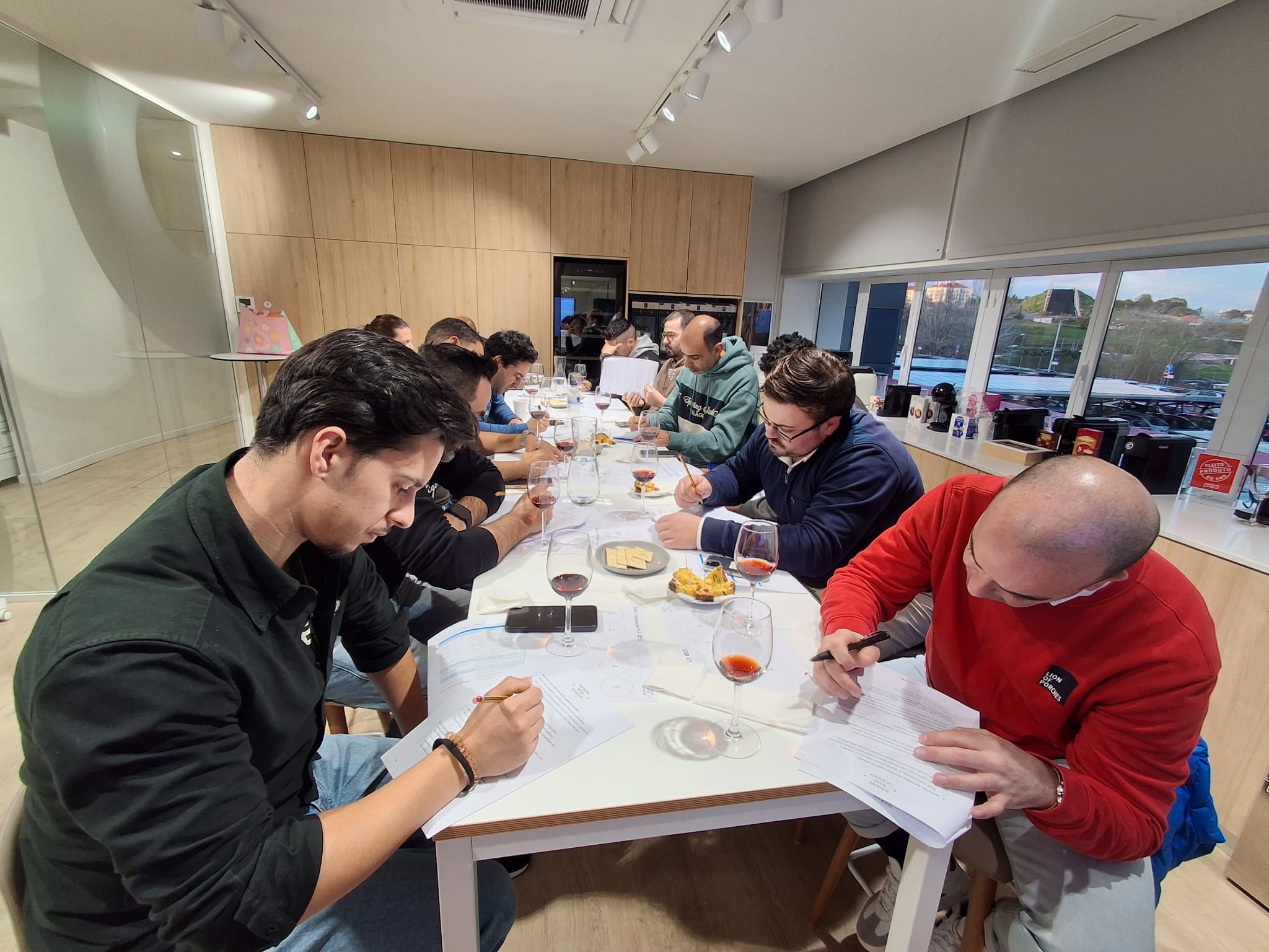 Um grupo de pessoas está sentado em torno de uma mesa branca, degustando bebidas e anotando em papéis, num ambiente de sala de provas moderna