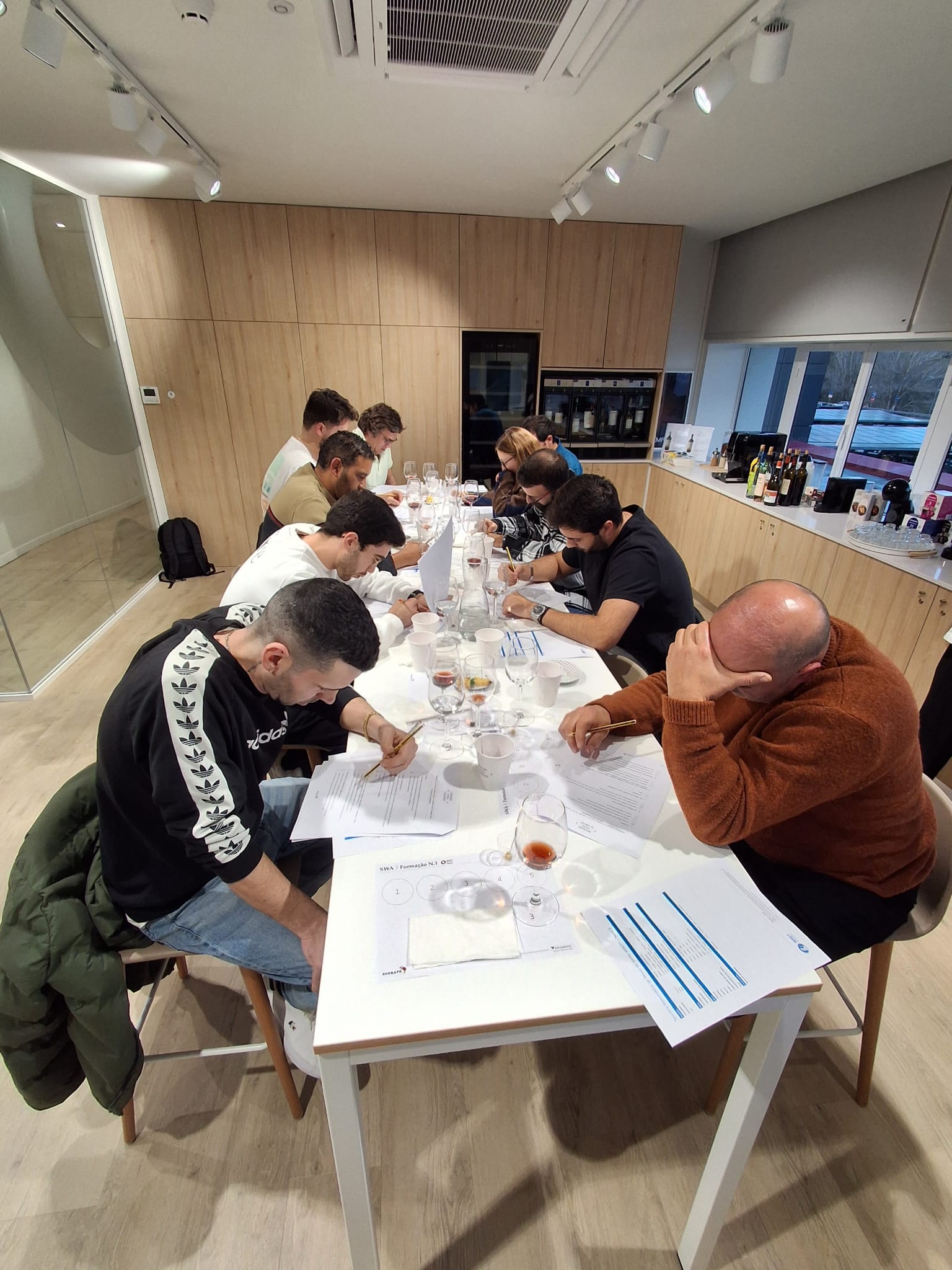 Um grupo de pessoas está focado a escrever em papéis dispostos sobre uma mesa com vários copos de degustação, numa sala moderna com janelas que revelam uma vista exterior
