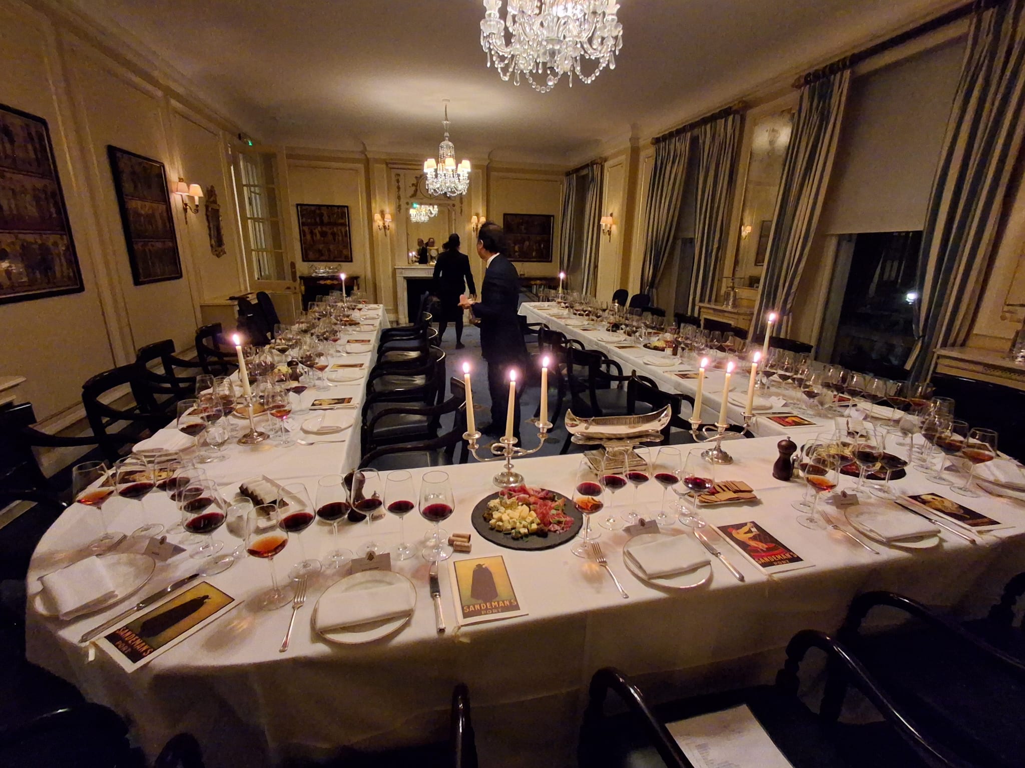 Uma sala de jantar elegante com uma mesa em formato de "U" posta com numerosos copos de vinho, velas, petiscos e materiais de degustação