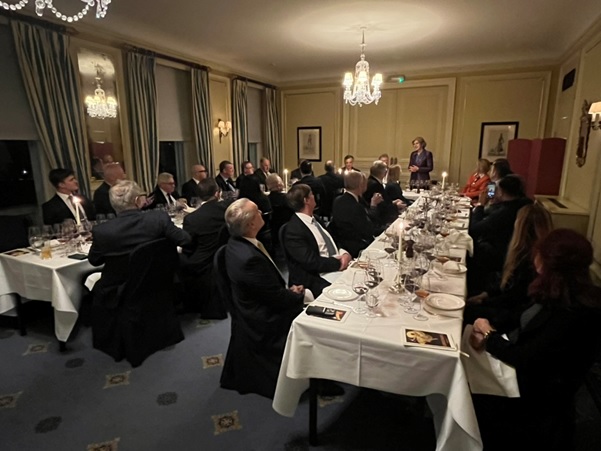 Uma mulher discursa para um grupo de pessoas sentadas formalmente à volta de uma mesa em "U" com velas e vários copos, num salão elegantemente decorado