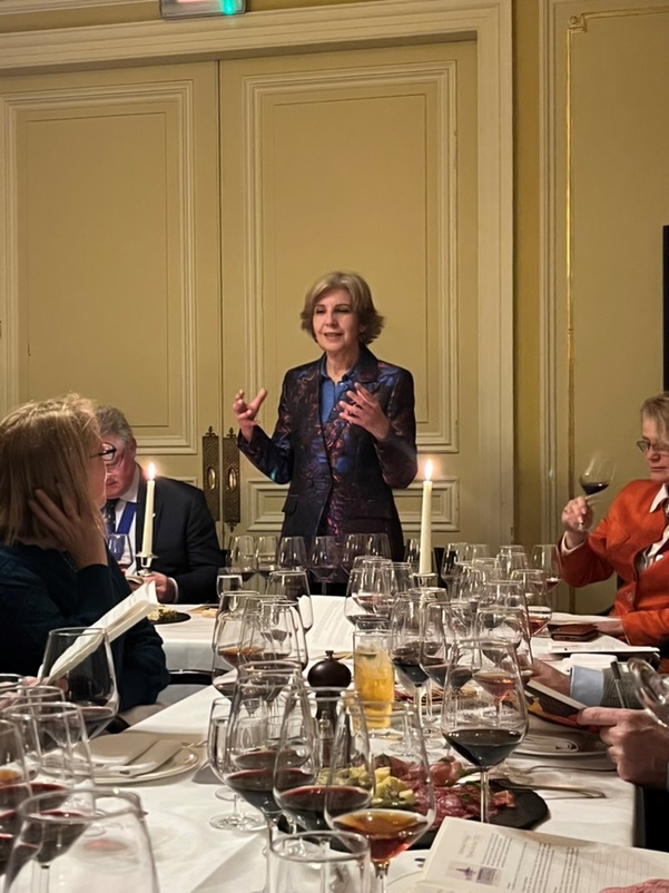 Uma mulher está a discursar para um grupo de pessoas sentadas formalmente à volta de uma mesa decorada com muitos copos de vinho e velas