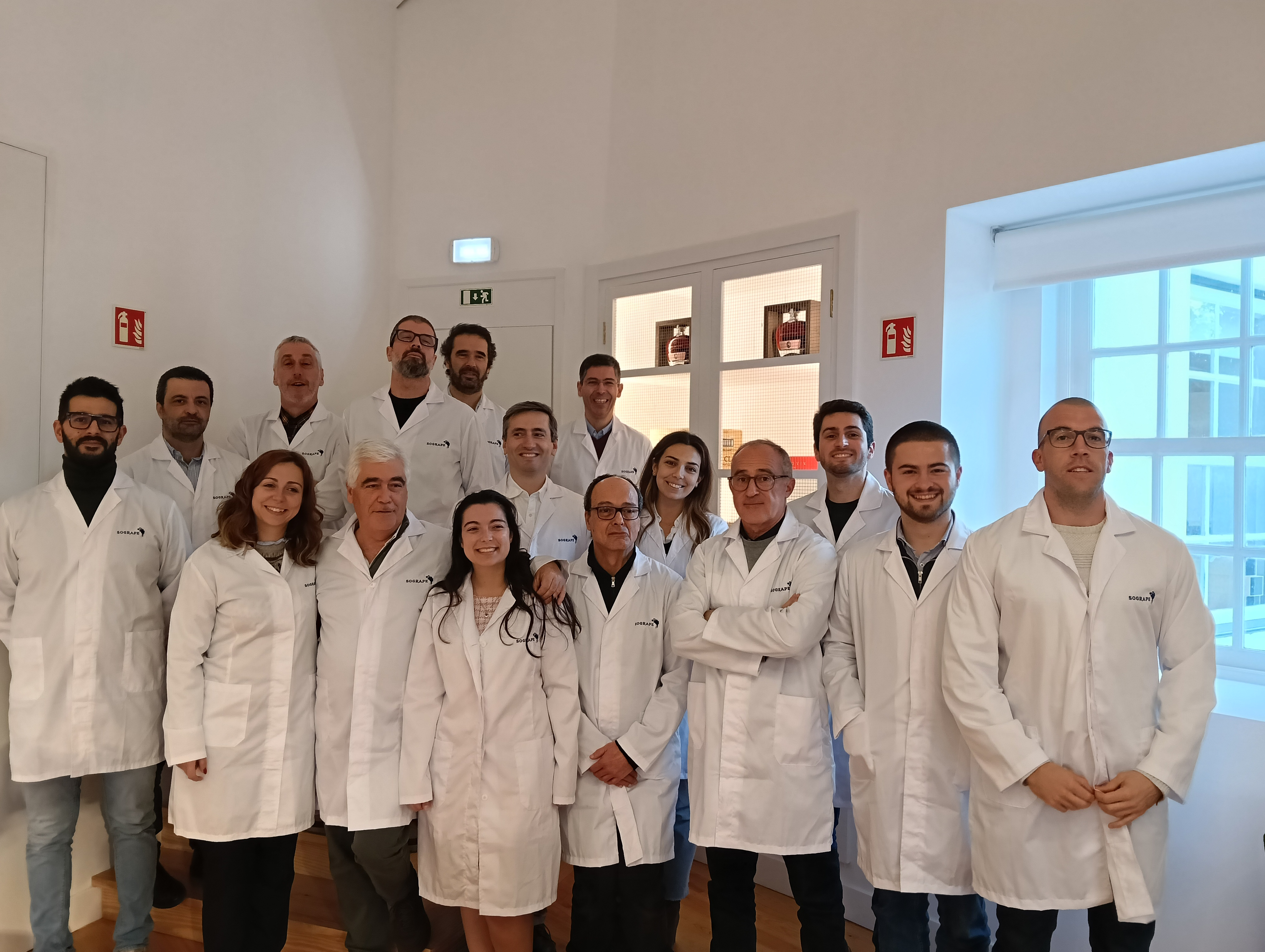 Um grupo de homens e mulheres, a maioria usando batas brancas, posa para uma fotografia num interior moderno e bem iluminado