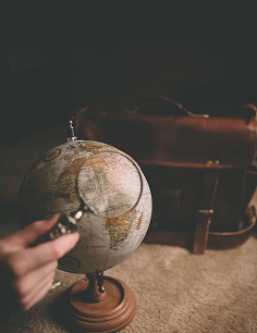Uma mão segura uma lupa sobre um globo terrestre, com uma mala de viagem em segundo plano, sugerindo exploração ou estudo