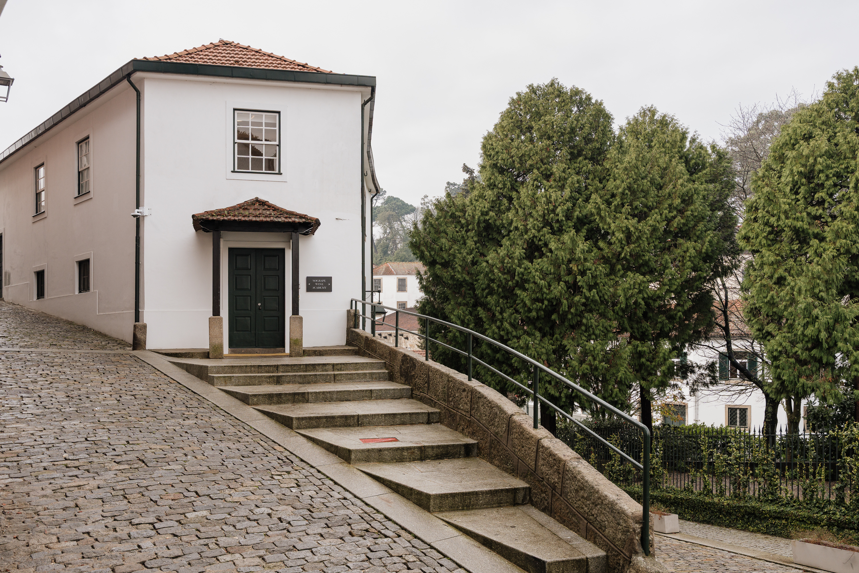 Um edifício branco com telhado de telha e uma escadaria de pedra, ladeado por árvores verdes e uma rua de calçada