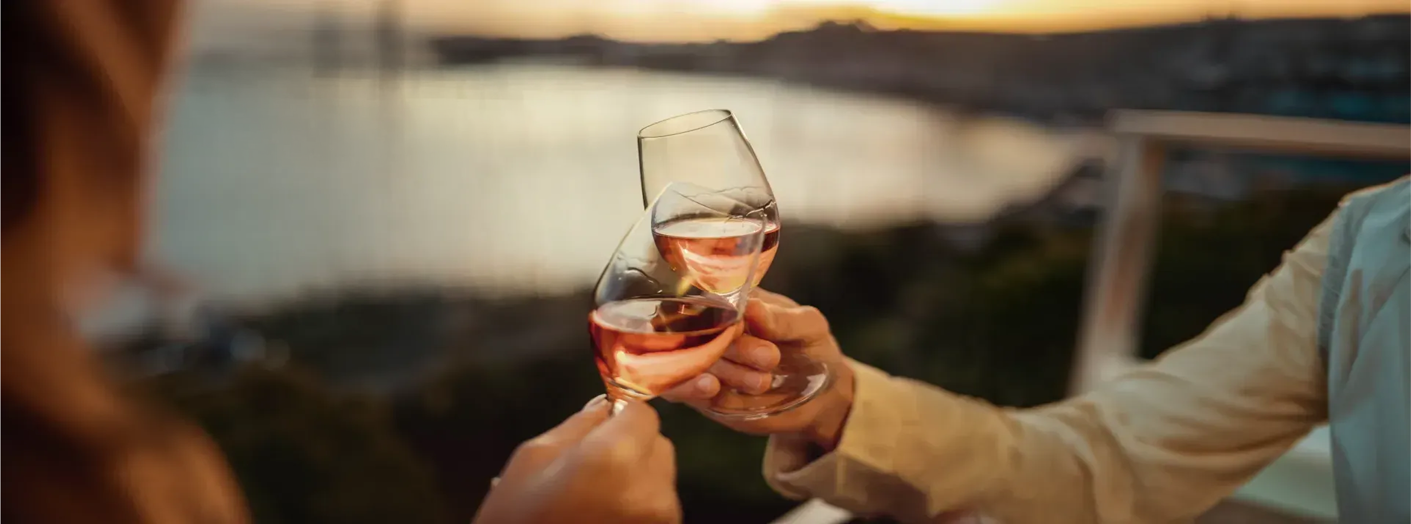 Duas mãos brindam com copos de vinho rosé ao pôr do sol, com um fundo desfocado de água e paisagem
