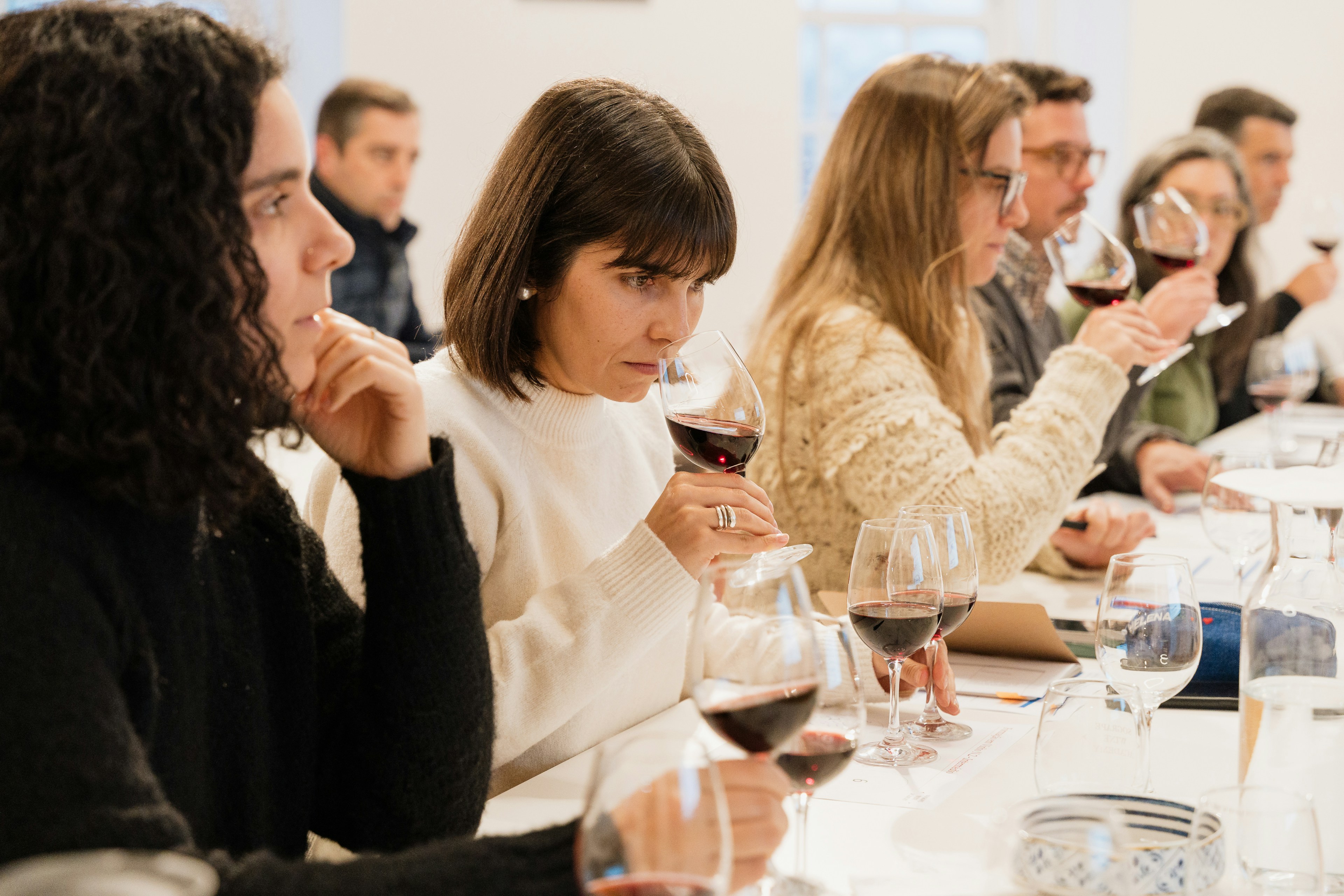Pessoas participam numa prova de vinhos, segurando e cheirando copos de vinho tinto à mesa
