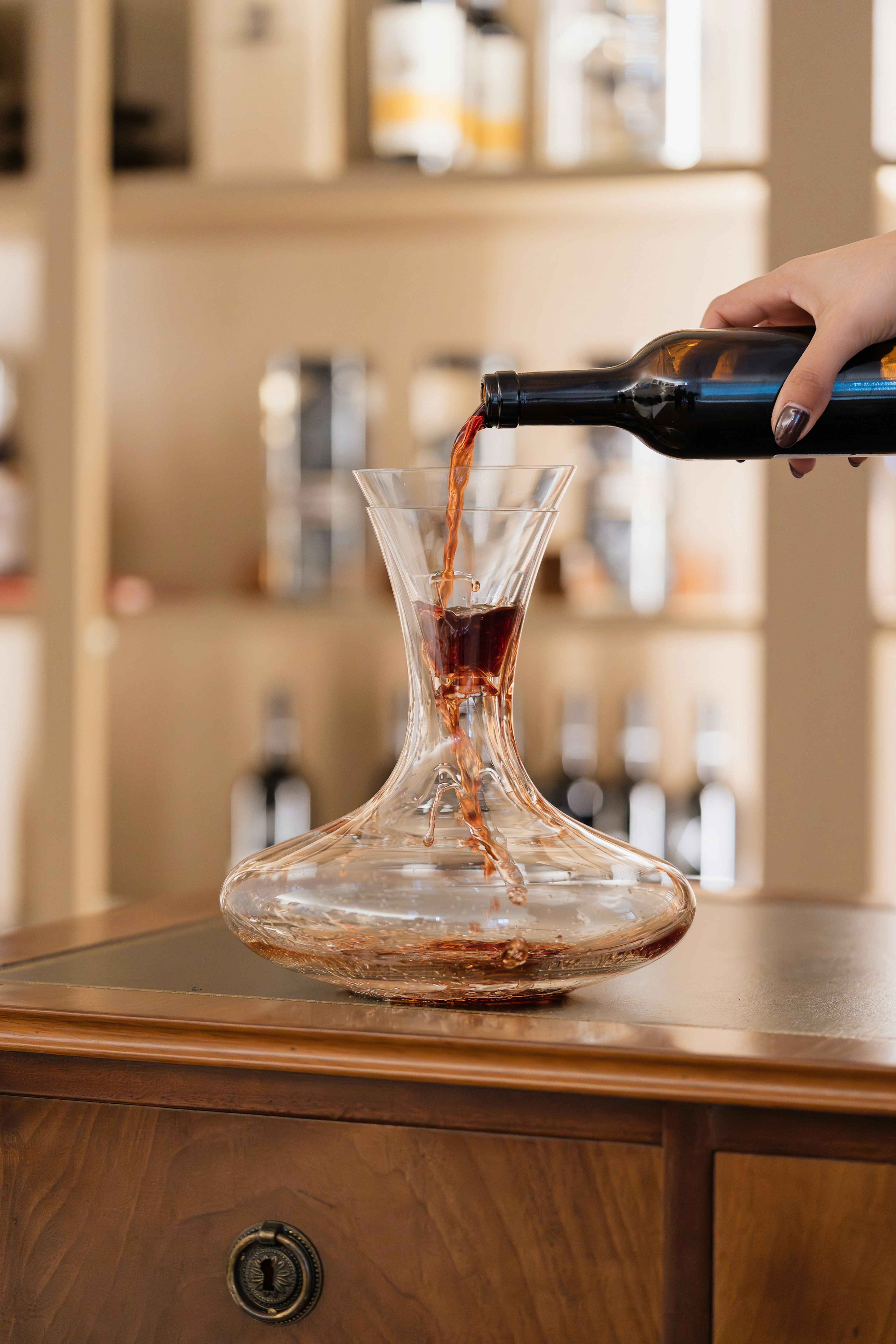 Uma mão verte vinho tinto de uma garrafa para uma garrafeira de vidro, sobre um móvel de madeira, com garrafas em prateleiras ao fundo