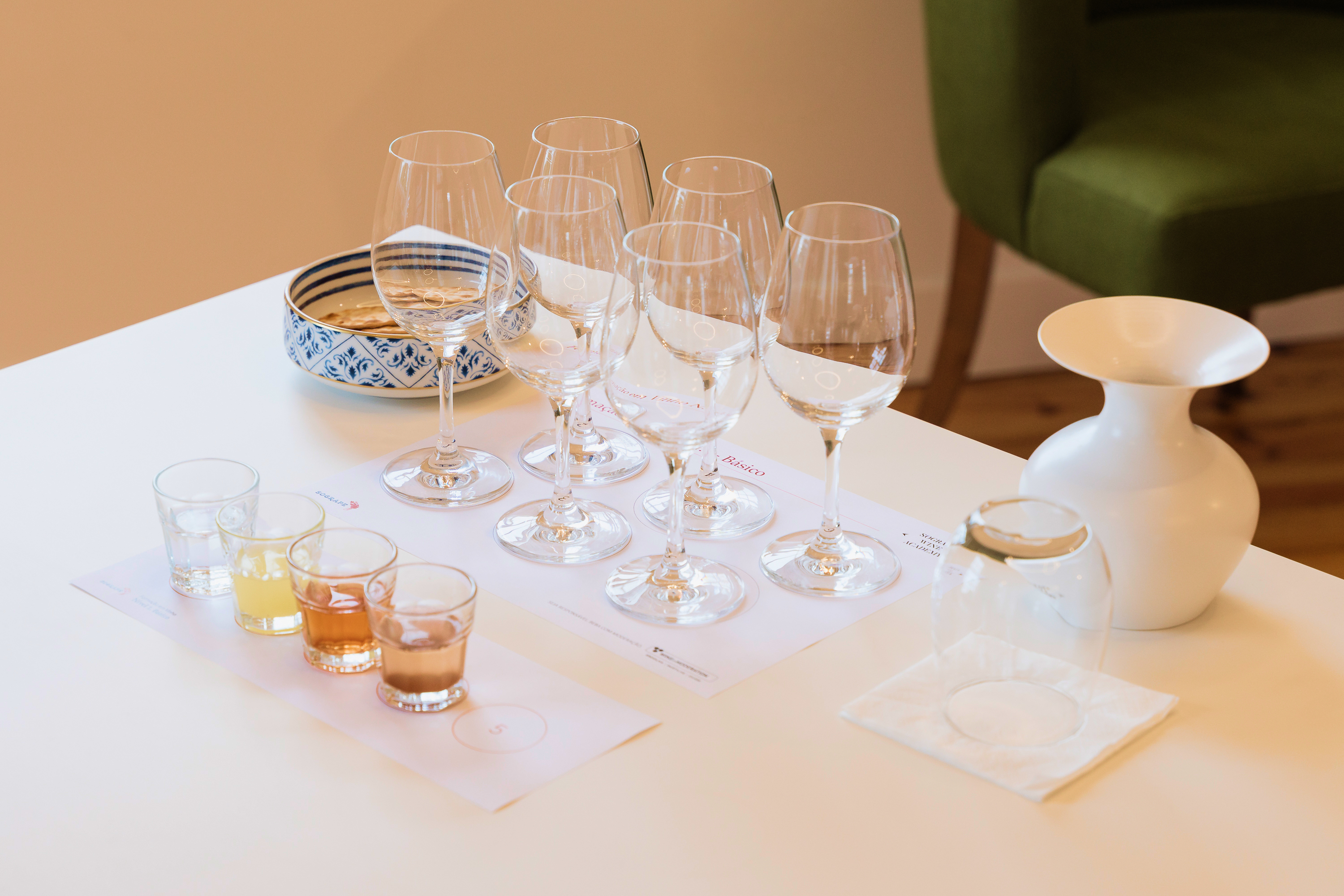 Uma mesa branca está posta para uma prova, com copos de vinho vazios, pequenos copos com líquidos coloridos, e um recipiente com snacks, num ambiente de sala de formação