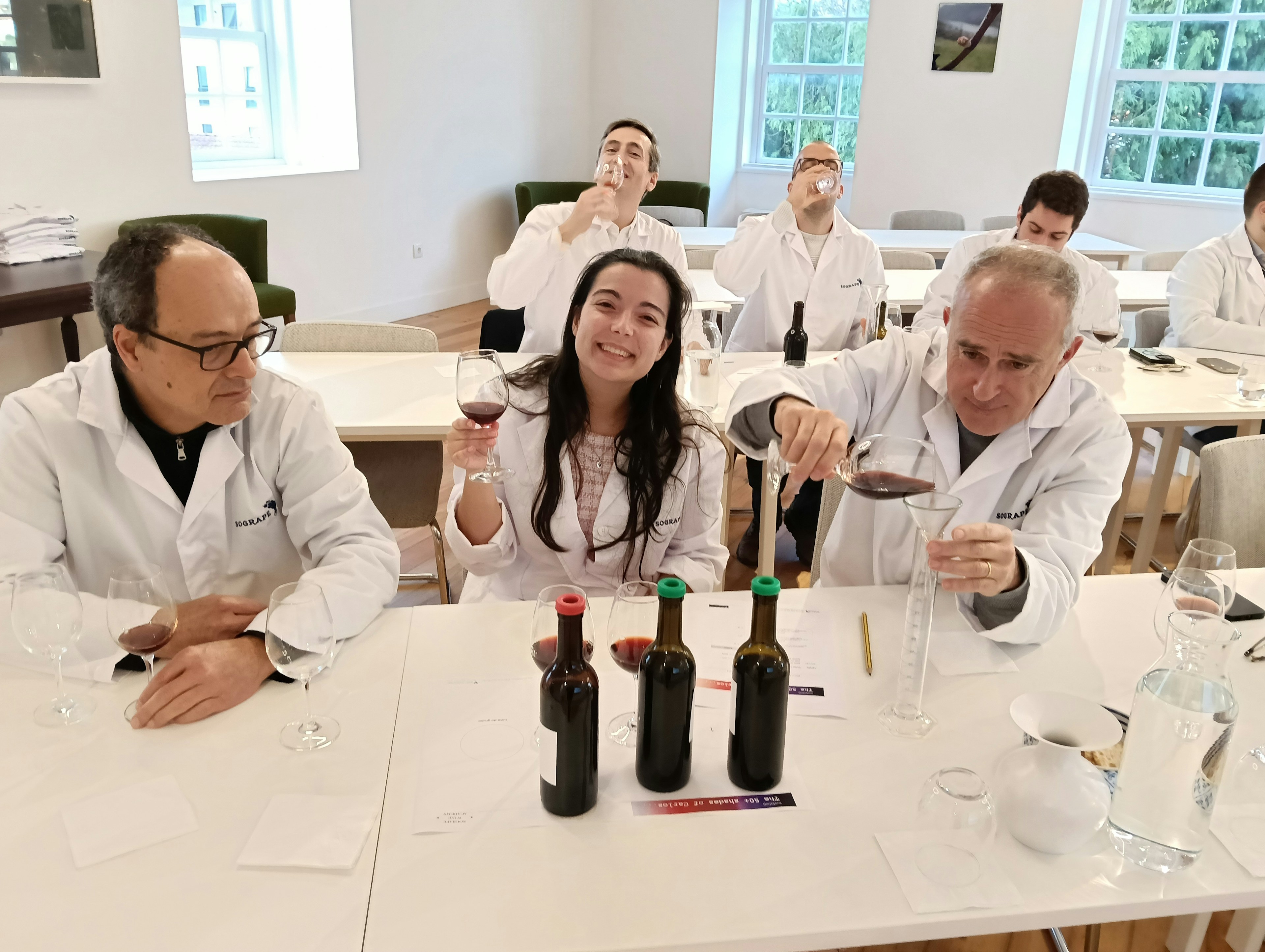 Um grupo de pessoas com batas brancas participa numa degustação de vinhos, alguns a servir e outros a provar, num ambiente de sala de provas iluminada