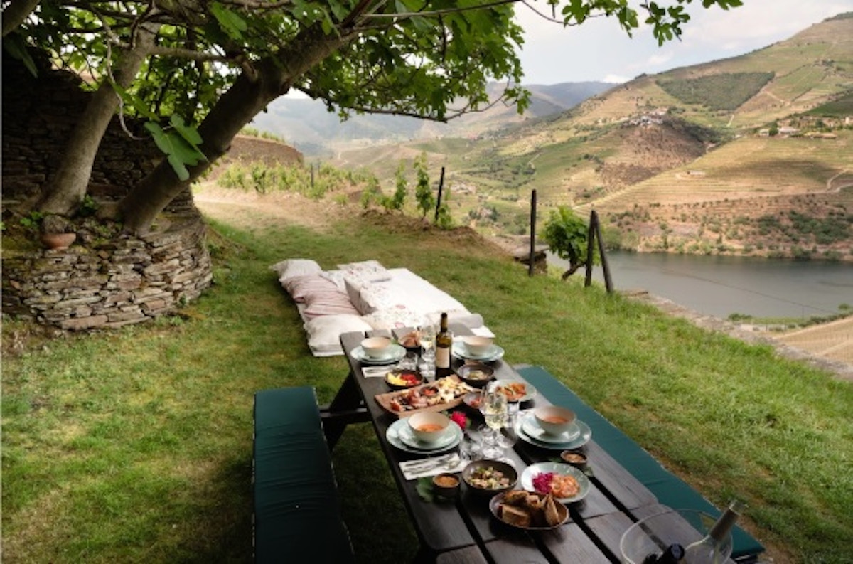 Uma mesa de piquenique com comida e bebida está posta num relvado junto a um rio, com colinas de vinhedos em socalcos ao fundo, sob um céu claro