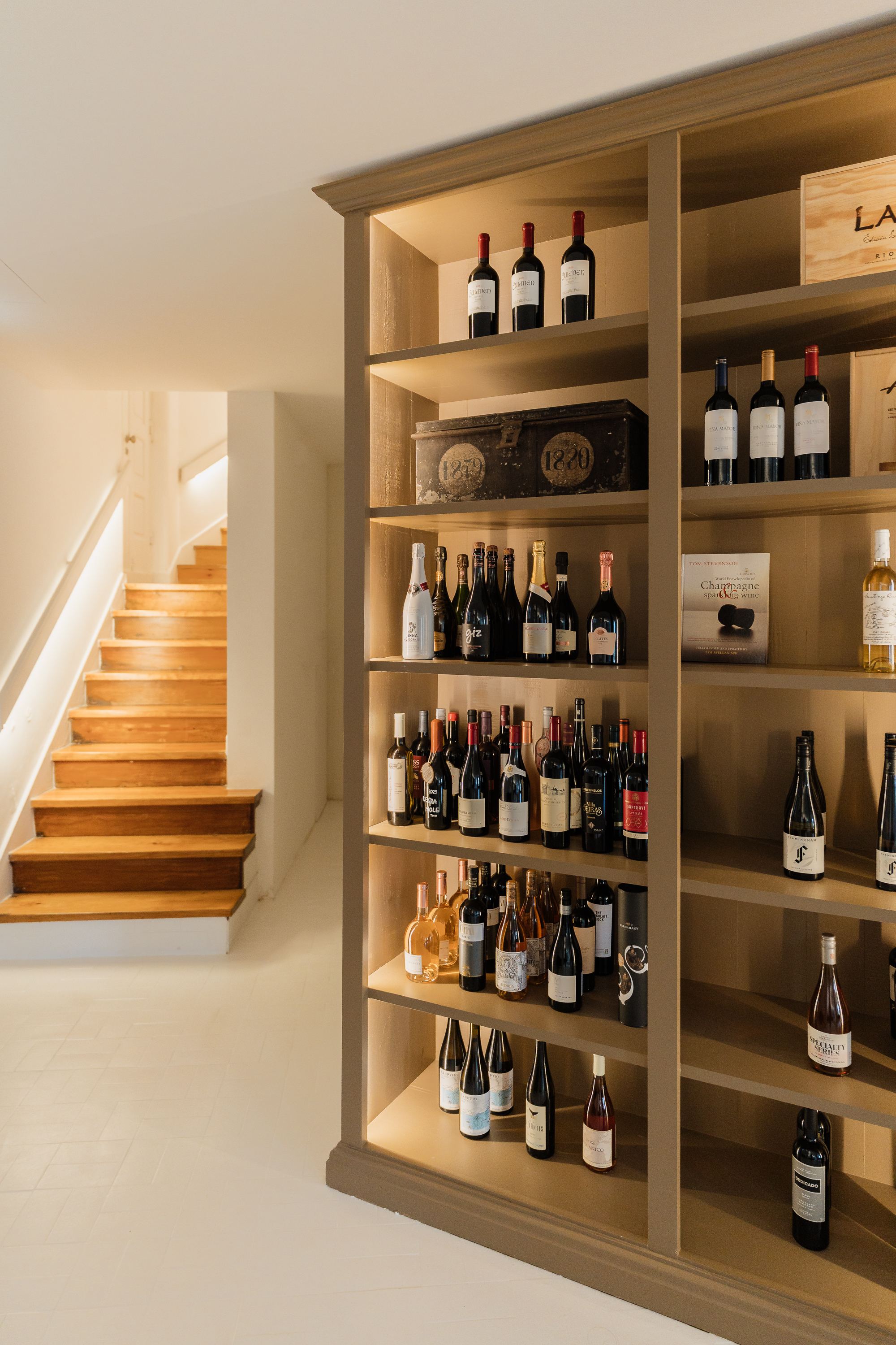 Uma vasta coleção de garrafas de vinho é exibida em prateleiras iluminadas, ao lado de uma escadaria de madeira