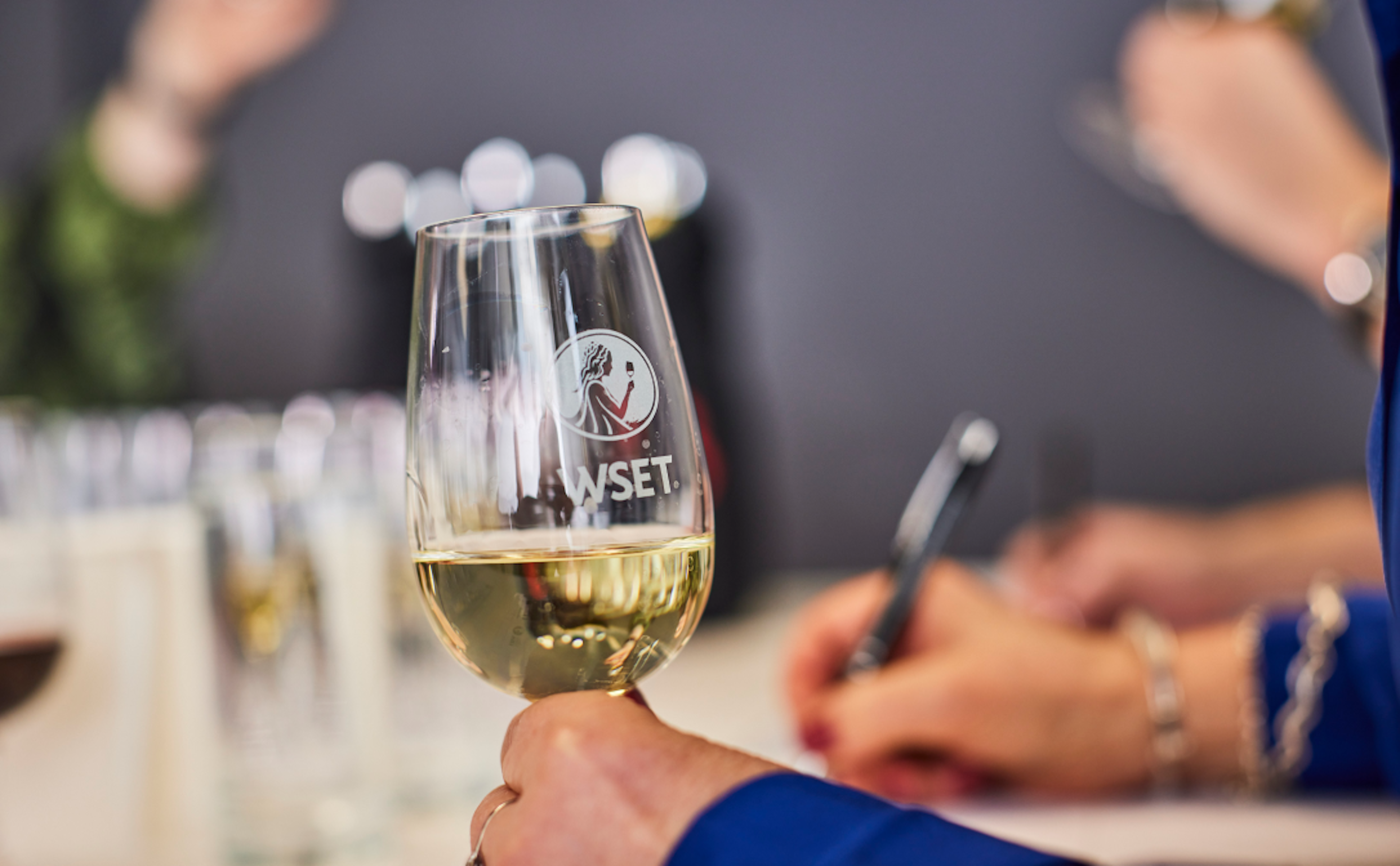Uma mão segura uma taça de vinho branco com o logótipo WSET, enquanto outra pessoa escreve no fundo, sugerindo uma aula ou prova de vinhos