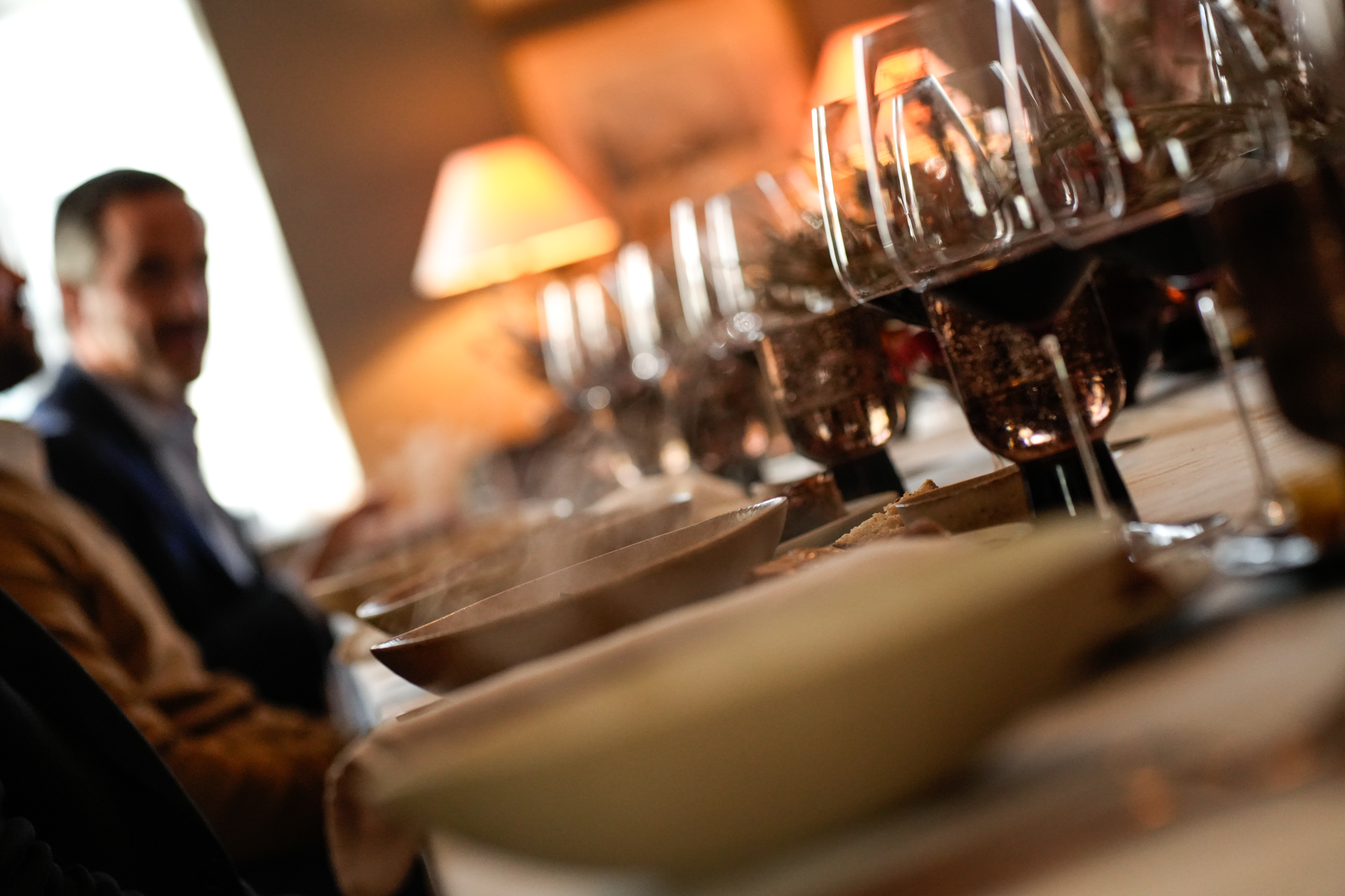 Uma mesa está posta com várias tigelas a vapor e uma fila de copos de vinho, com uma pessoa desfocada ao fundo, criando um ambiente acolhedor