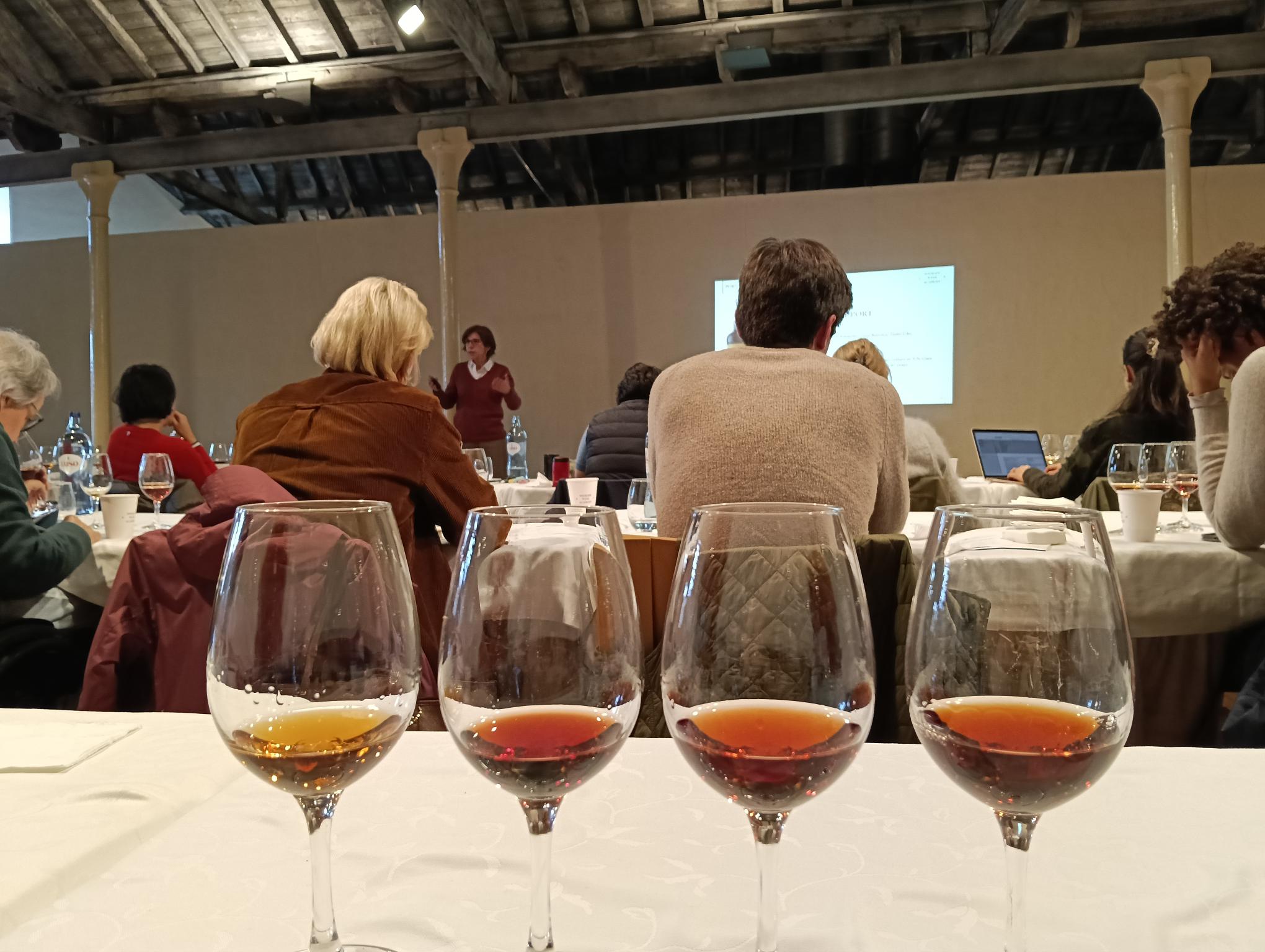 Quatro copos de vinho com diferentes tonalidades estão em primeiro plano numa mesa, enquanto um grupo de pessoas assiste a uma apresentação ao fundo