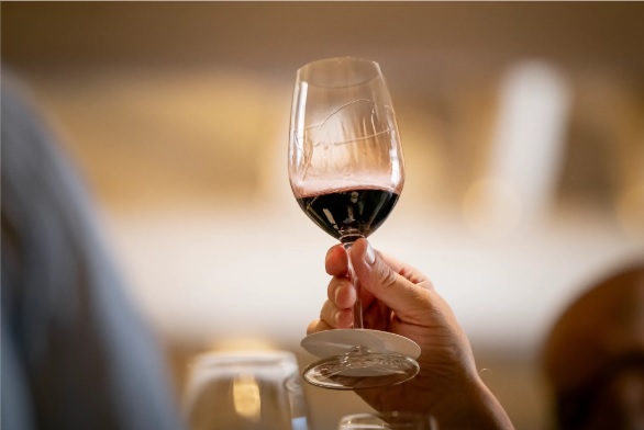 Uma mão segura um copo de vinho tinto, com as "lágrimas" do vinho visíveis no vidro, num ambiente com luz difusa