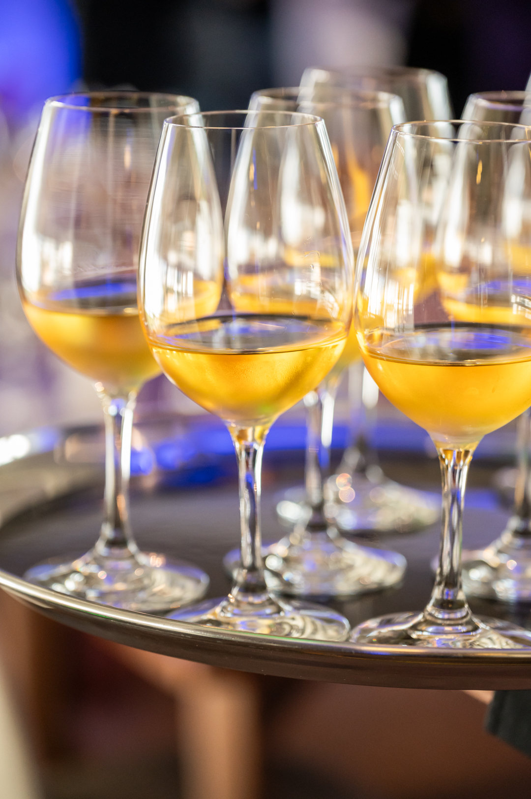 Uma bandeja com vários copos de vinho, a maioria cheios de uma bebida dourada, está a ser transportada