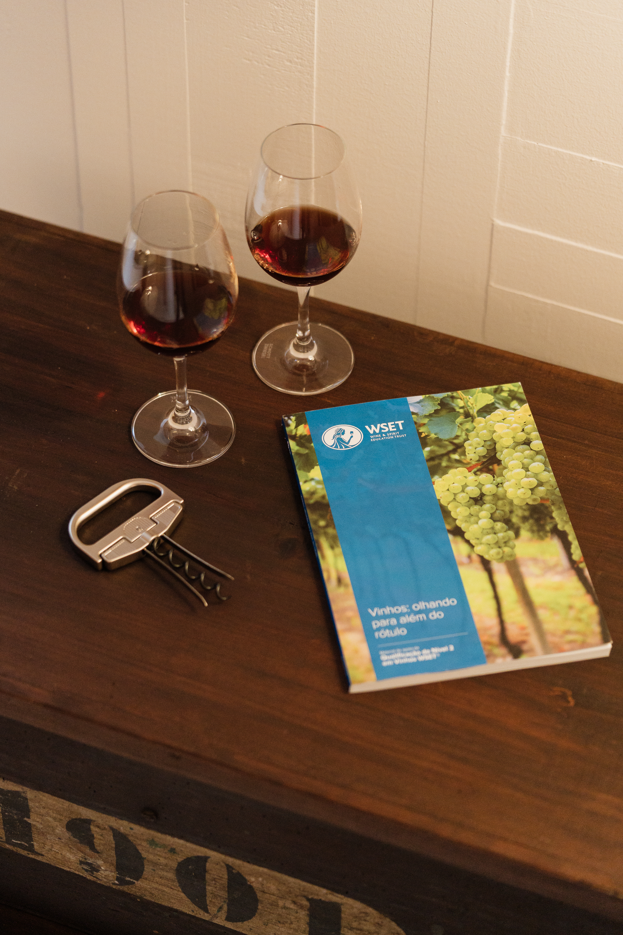 Uma superfície de madeira exibe dois copos de vinho tinto, um saca-rolhas e um manual WSET sobre vinhos