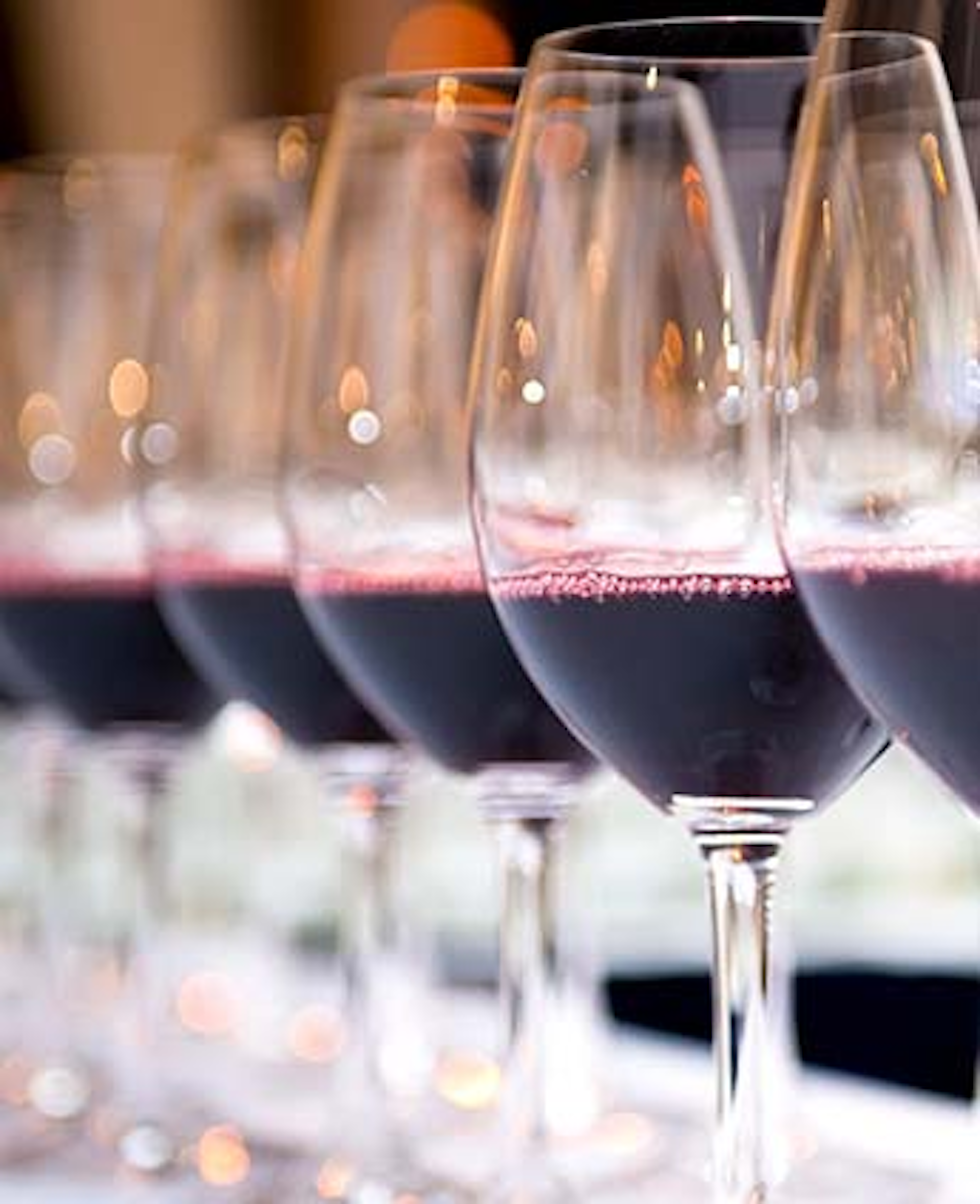 Uma fila de taças de vinho tinto, parcialmente preenchidas, está alinhada, com um fundo desfocado de luzes