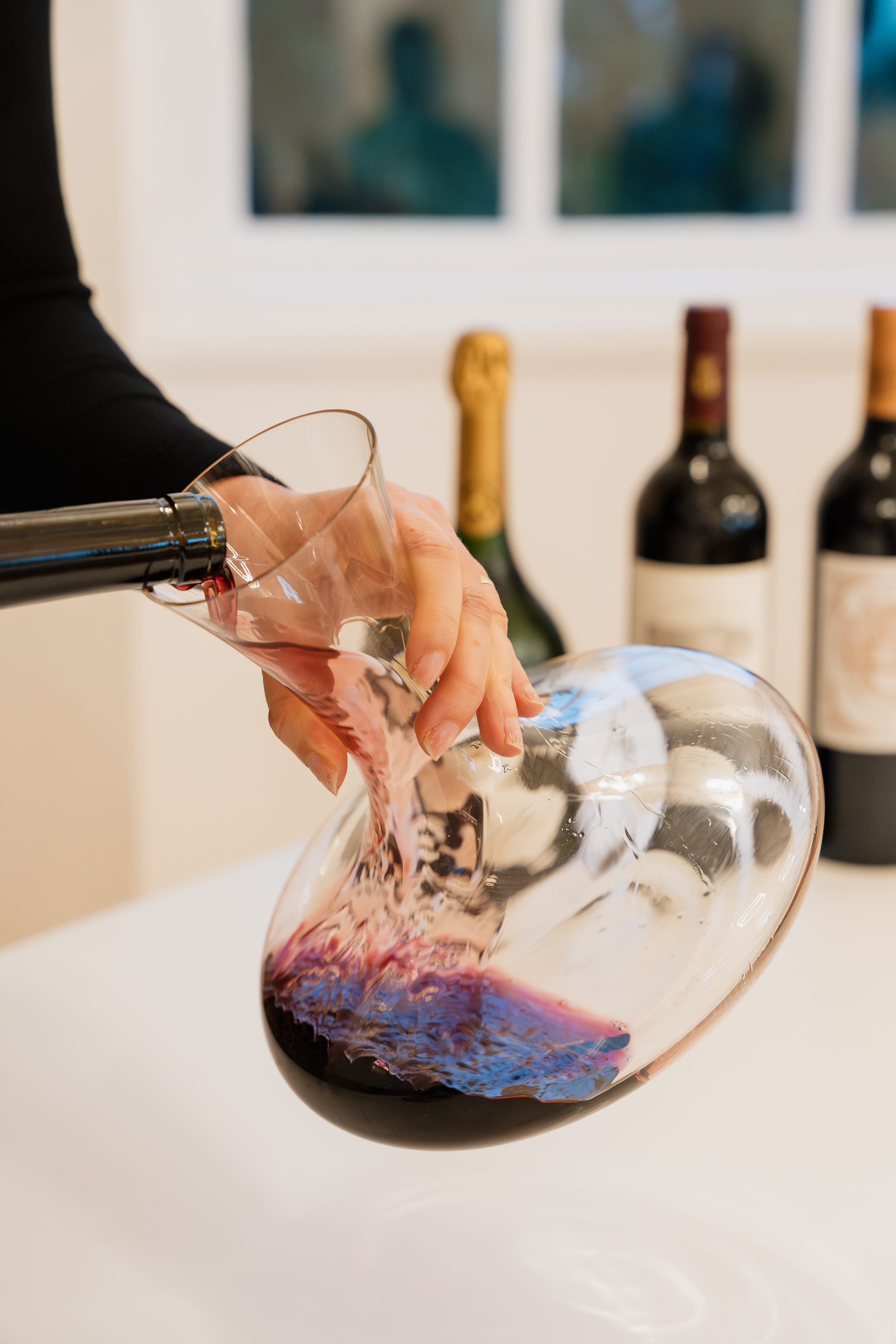 imagem mostra uma pessoa a servir vinho tinto para um decantador de vidro, com garrafas ao fundo