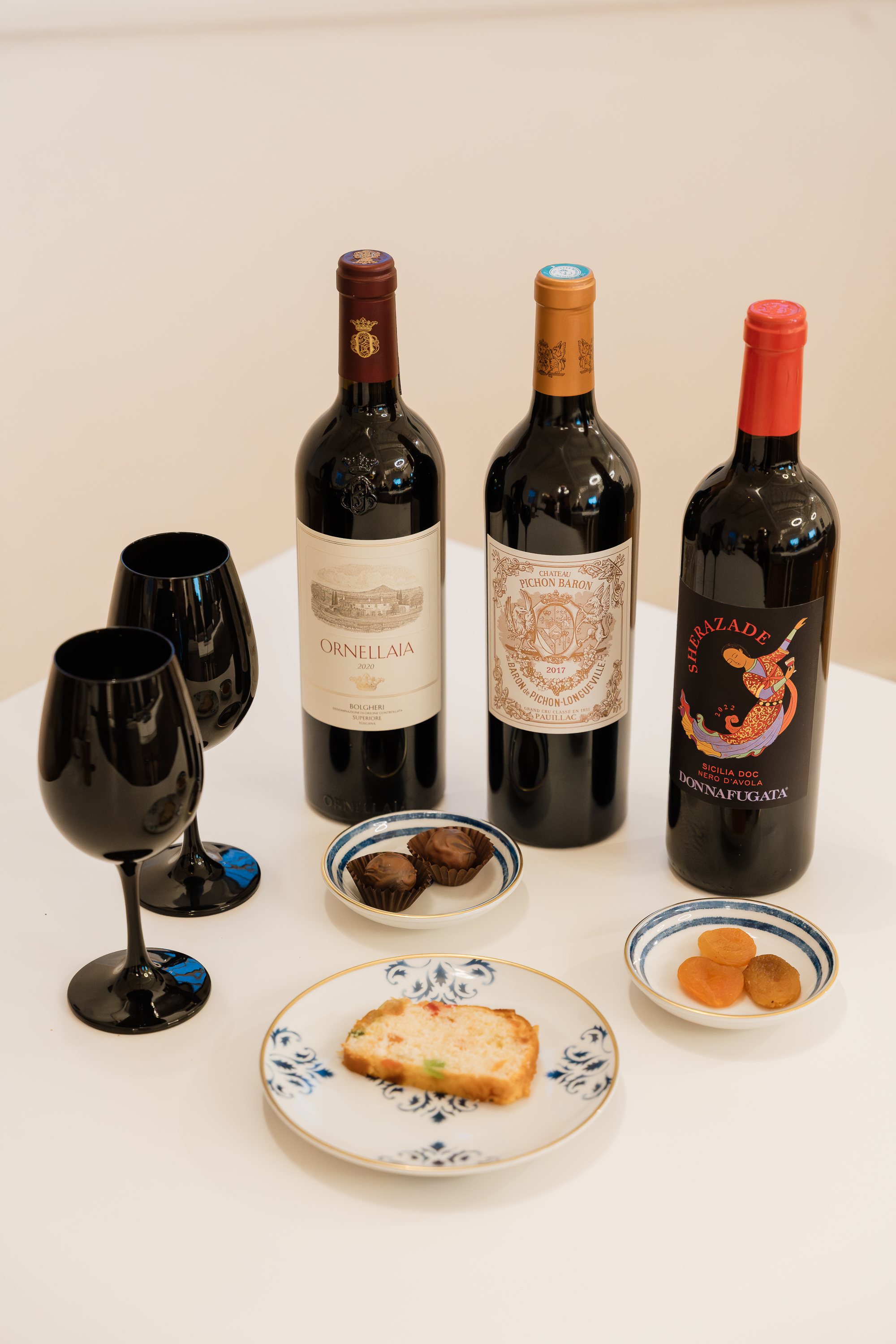 três garrafas de vinho numa mesa, com copos pretos e pequenos petiscos em pratos