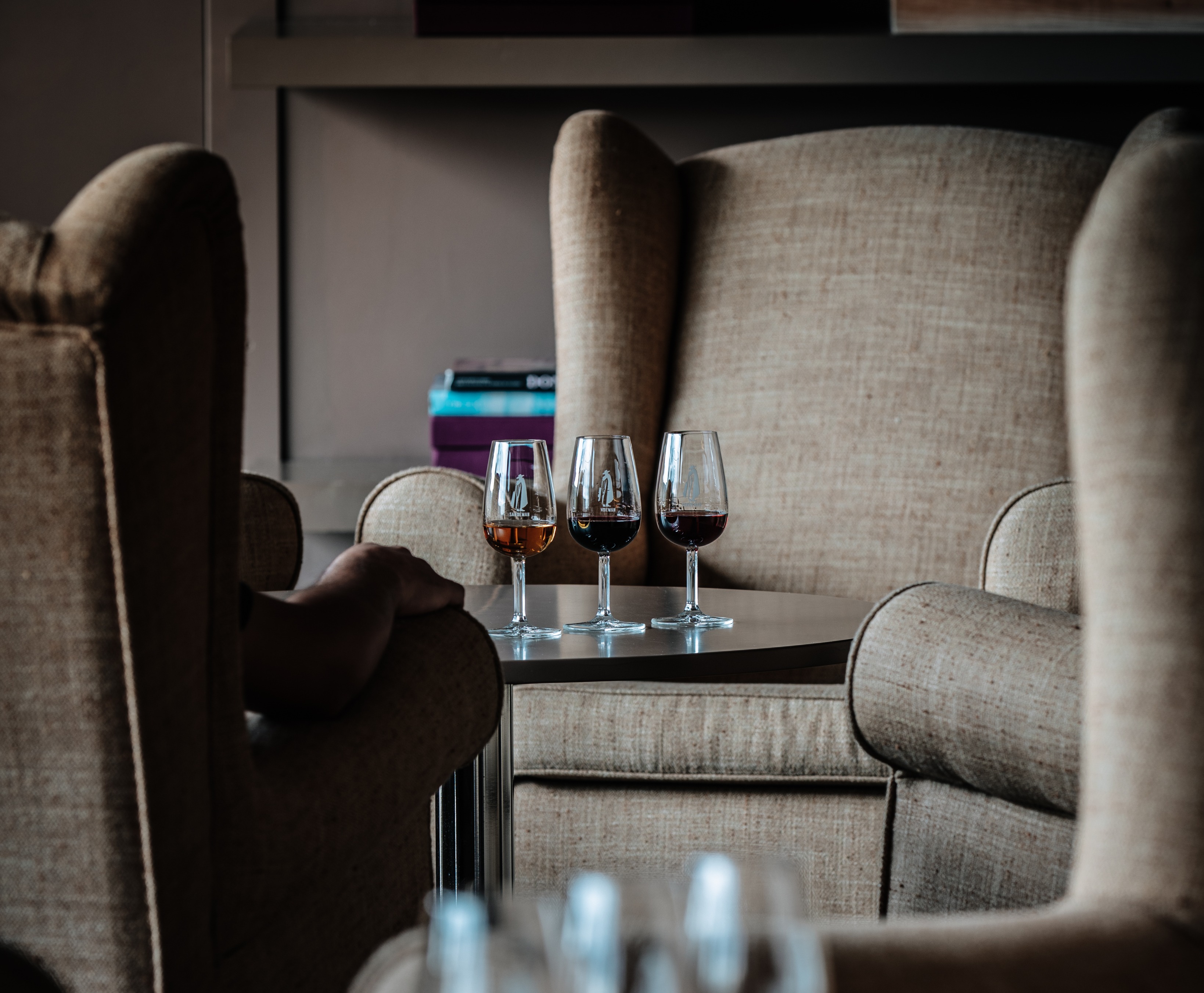 Três copos de vinho, com diferentes tonalidades, estão sobre uma mesa entre duas poltronas numa sala de estar