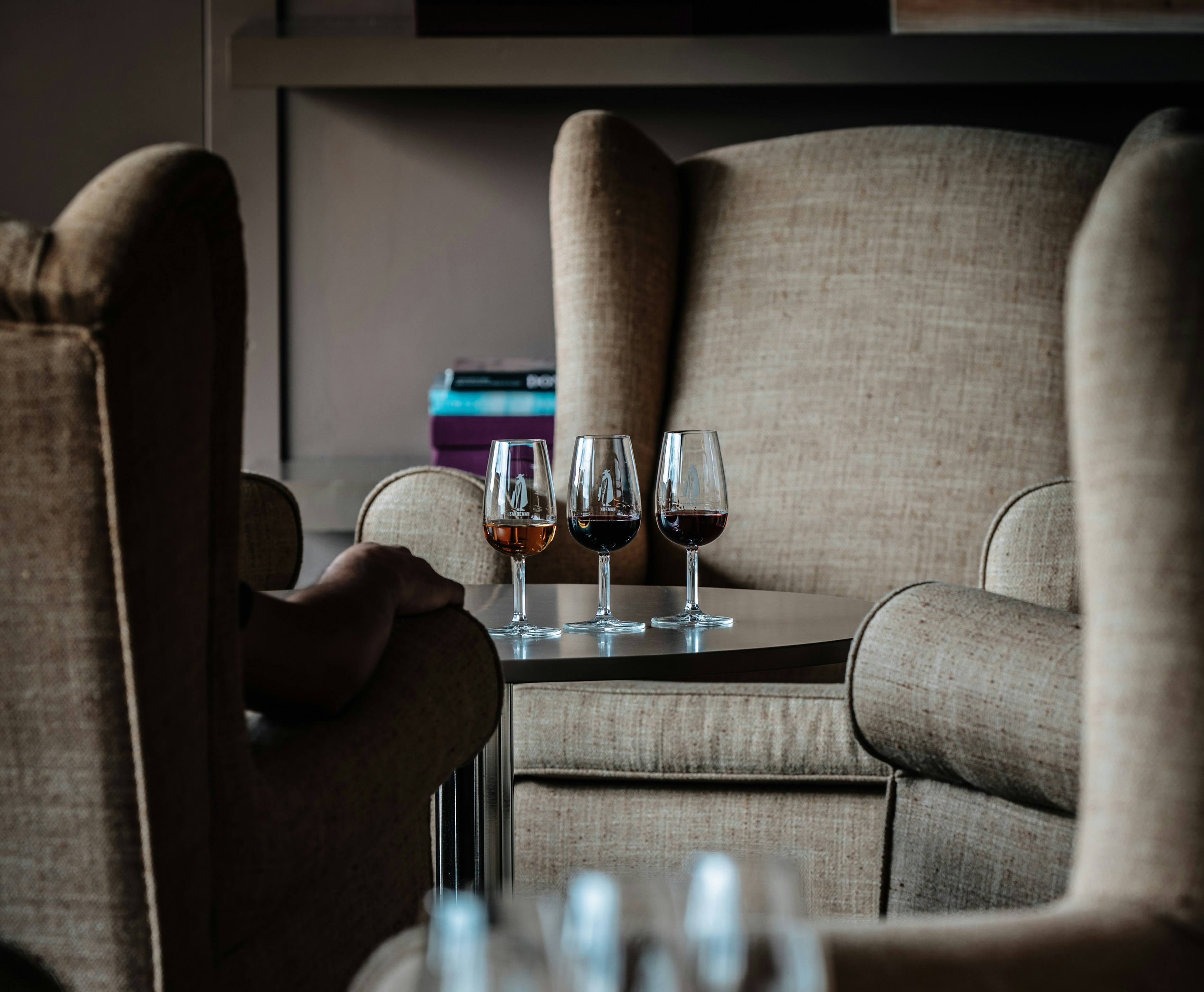 Três copos de vinho, com diferentes tonalidades, estão sobre uma mesa entre duas poltronas numa sala de estar
