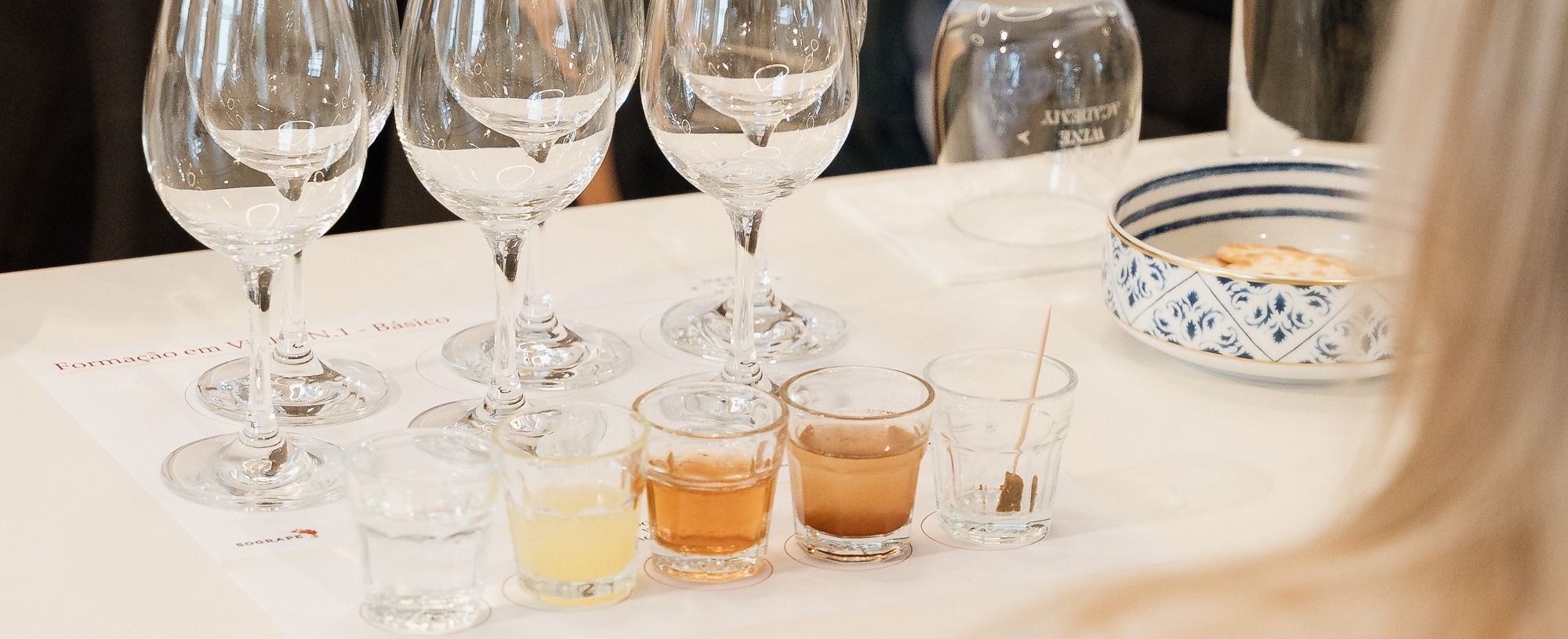 Uma mesa está preparada para uma prova ou formação, com várias taças de vinho, pequenos copos com líquidos coloridos e uma garrafa de água, com participantes visíveis em segundo plano