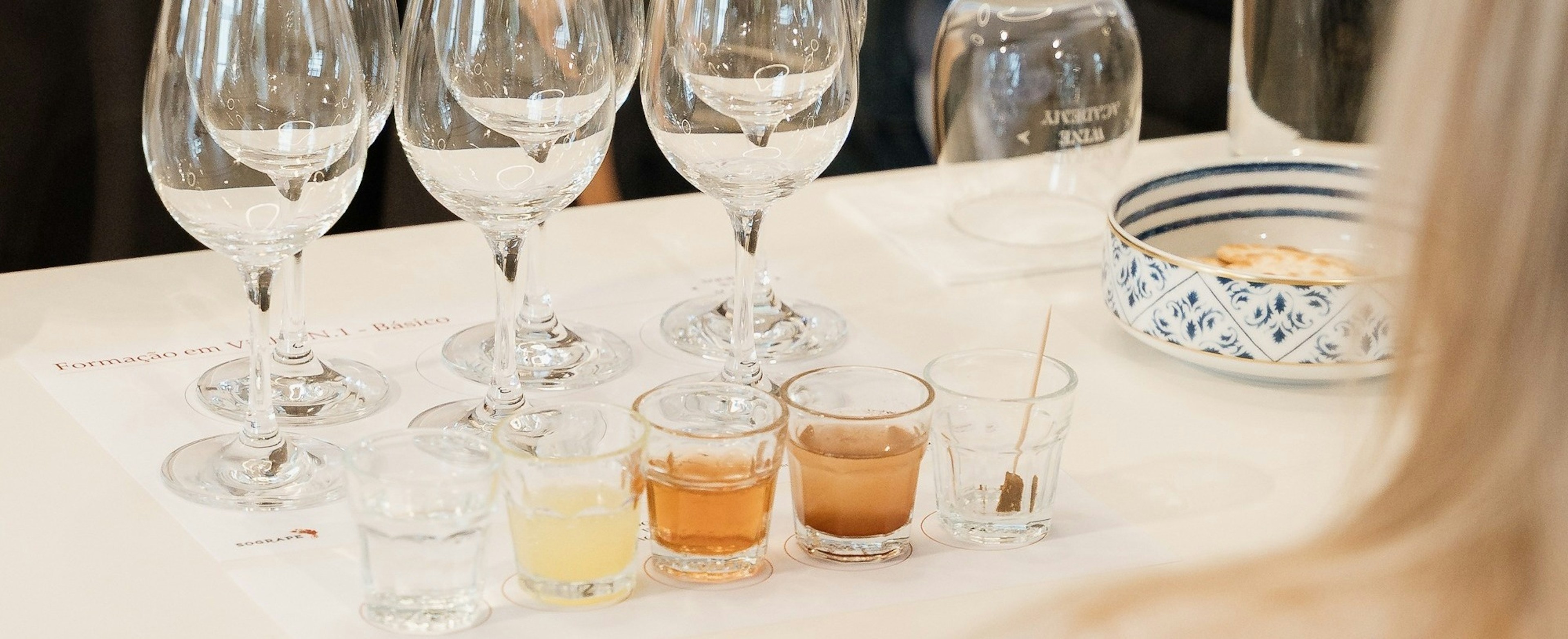 Uma mesa está preparada para uma prova ou formação, com várias taças de vinho, pequenos copos com líquidos coloridos e uma garrafa de água, com participantes visíveis em segundo plano