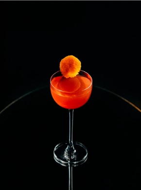 Um cocktail vermelho-alaranjado num copo de haste, adornado com uma bola de laranja, sobre um fundo escuro