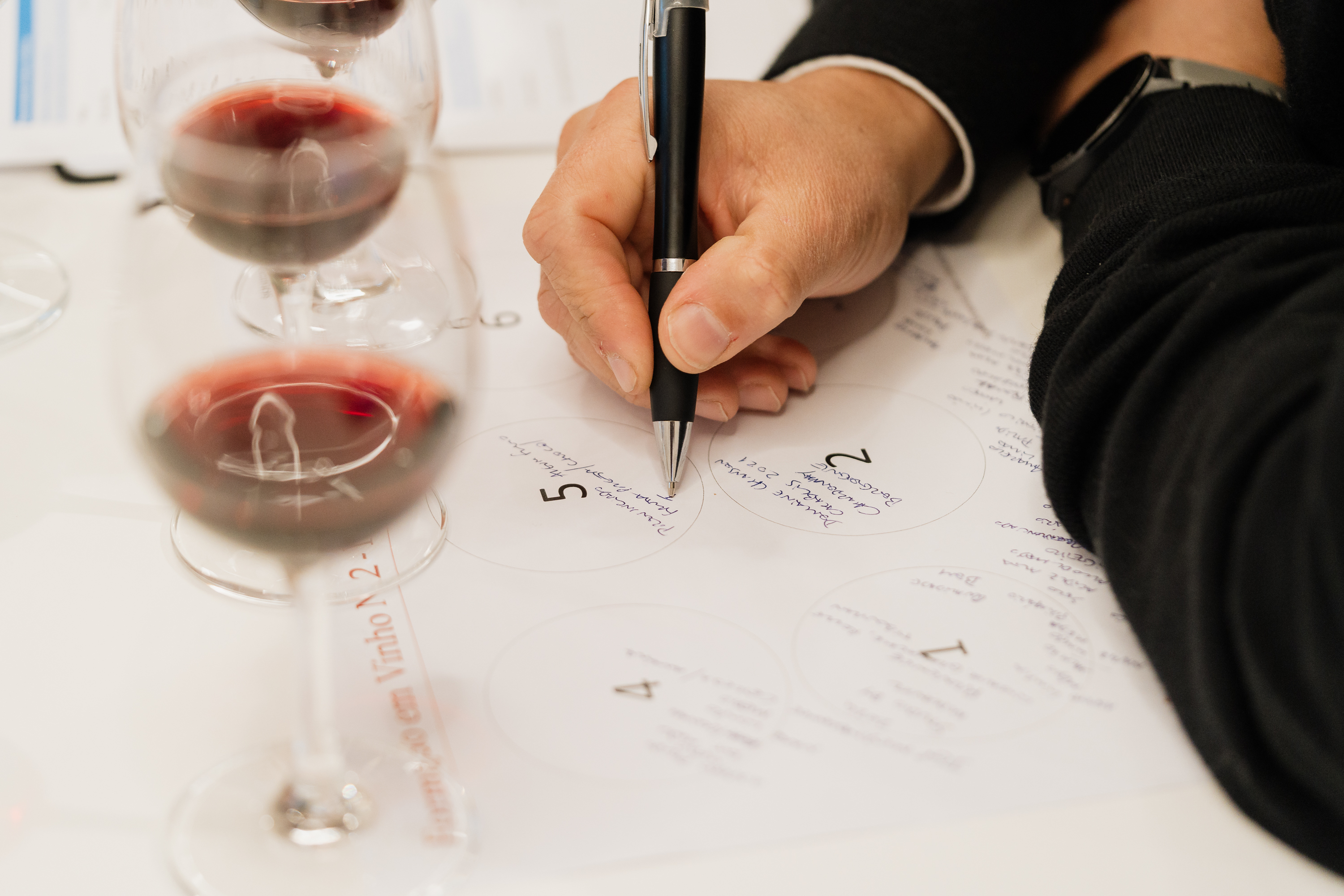 Uma pessoa escreve notas de prova num formulário com círculos numerados, com vários copos de vinho tinto em primeiro plano, alguns fora de foco