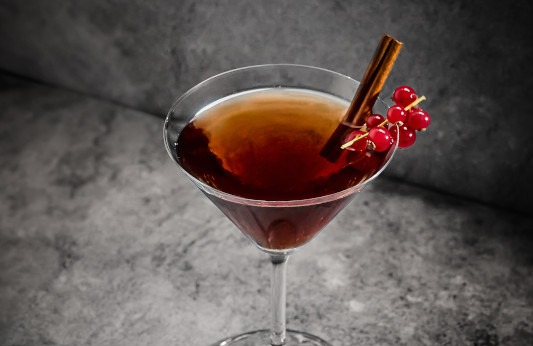 Um cocktail escuro num copo de martini, com um pau de canela e groselhas vermelhas, sobre um fundo cinzento escuro